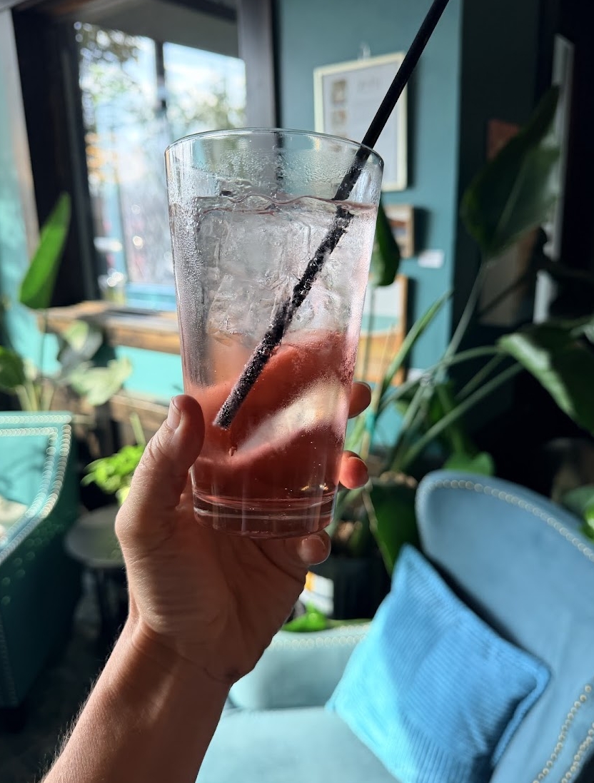 Hand holding a tall glass with a straw, ice, and a pink drink inside, in a room with plants and blue furniture.