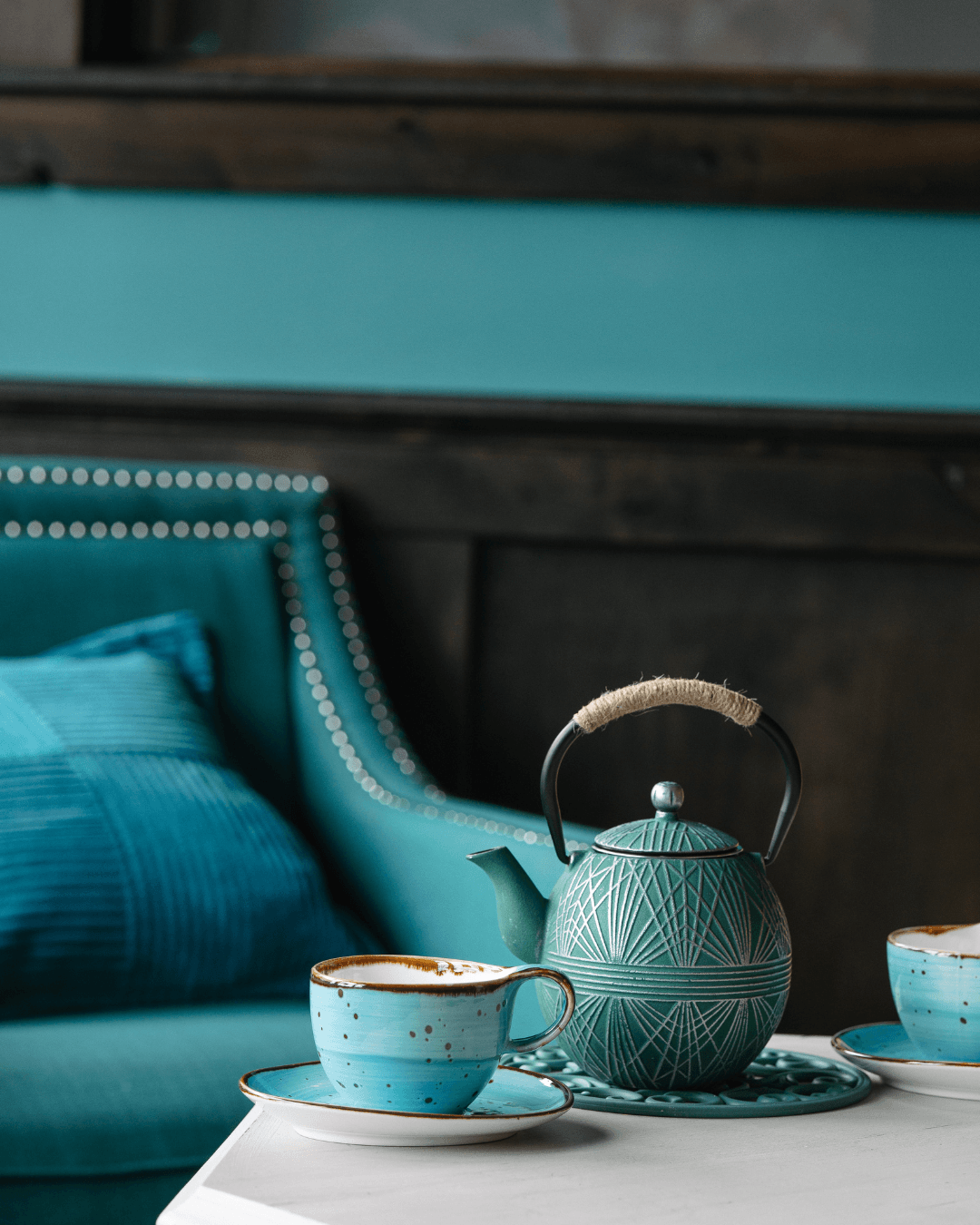 Teapot and cups on a table in front of a teal sofa with a pillow, in a cozy room setting.
