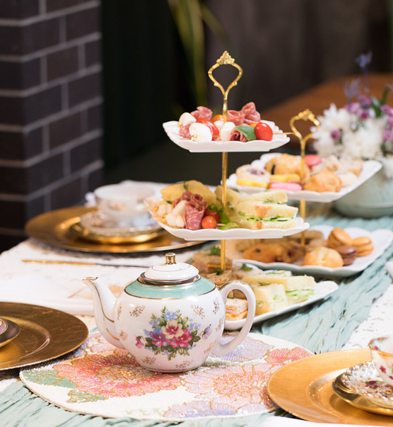Teapot with floral design on a table with a tiered tray of desserts and a floral centerpiece in the background.