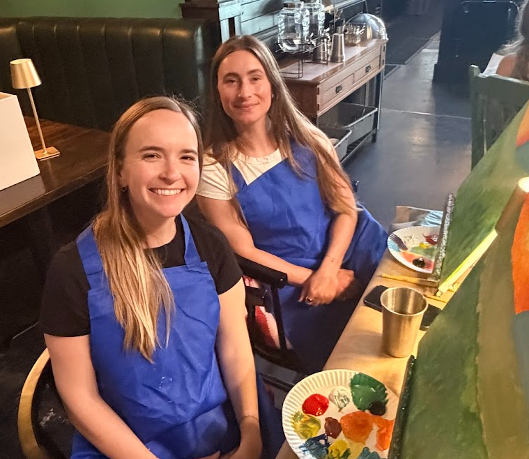 Two women sitting at a table with paint supplies, smiling, in a restaurant or cafe setting.