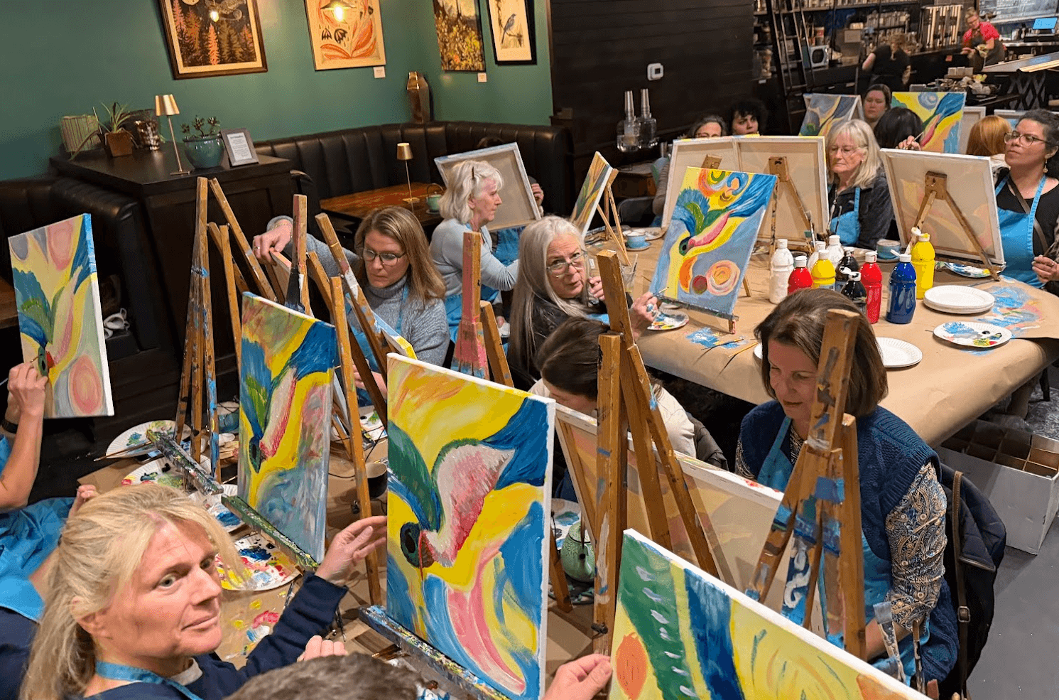 People painting colorful abstract artworks on canvases at a group art class in a room with teal walls.