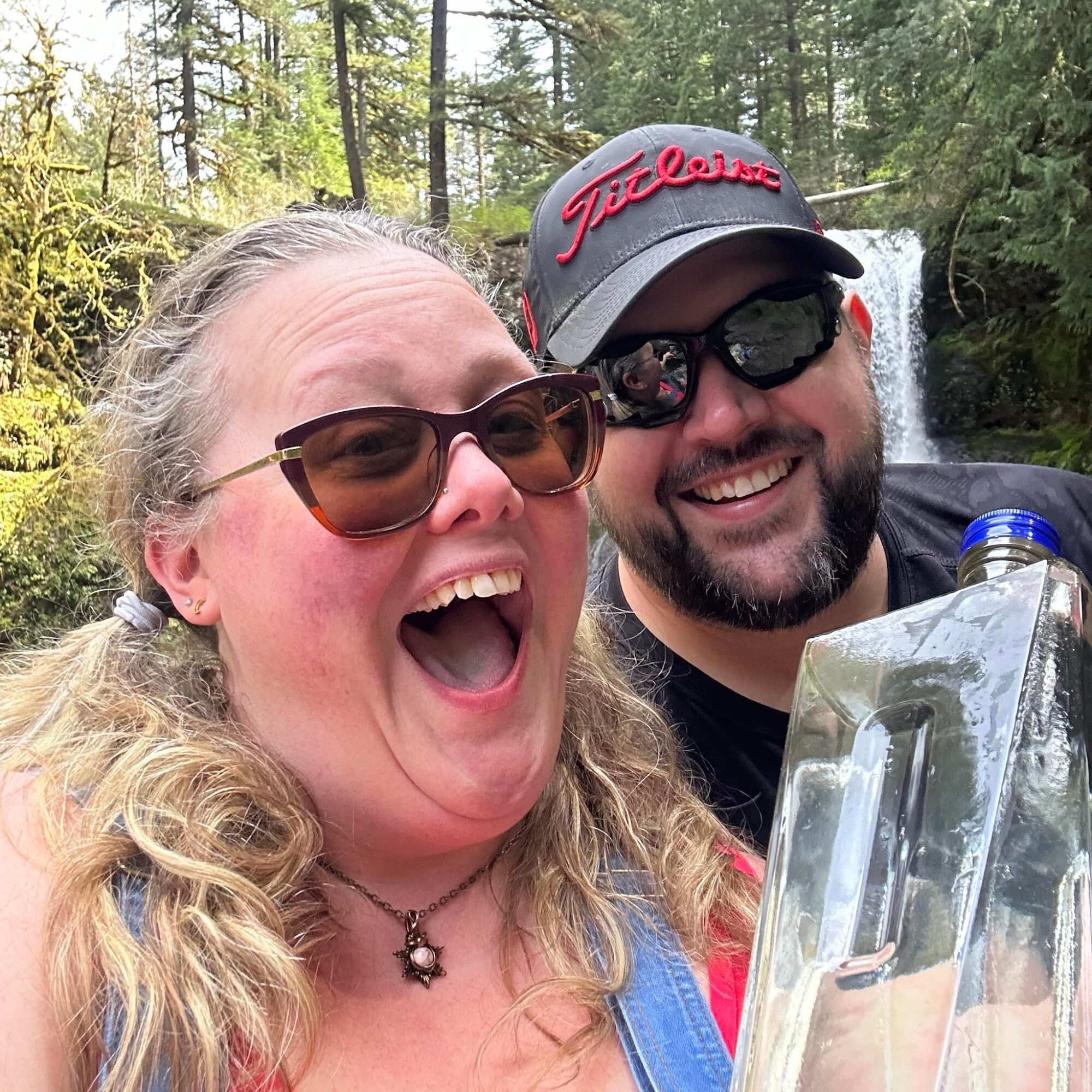 Two smiling people outdoors with trees and a waterfall in the background, one wearing sunglasses and a cap, the other with glasses and long hair.