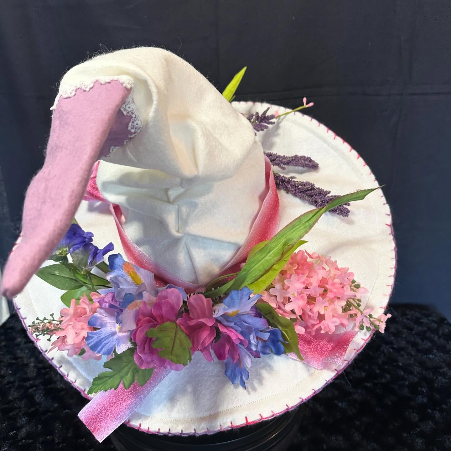 A decorative hat with a pink and white color scheme, adorned with flowers and a fabric bunny ear, on a black background.