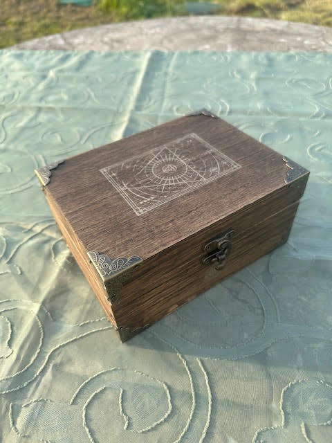 Wooden box with a metal latch on a textured fabric surface, top has a diagram or blueprint drawing, rectangular shape.