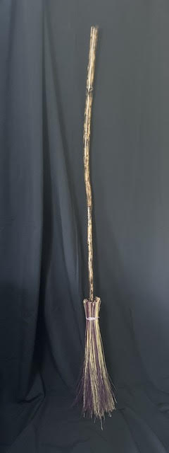 A long, thin broom with a handle and bristles at the bottom, standing upright against a dark background.