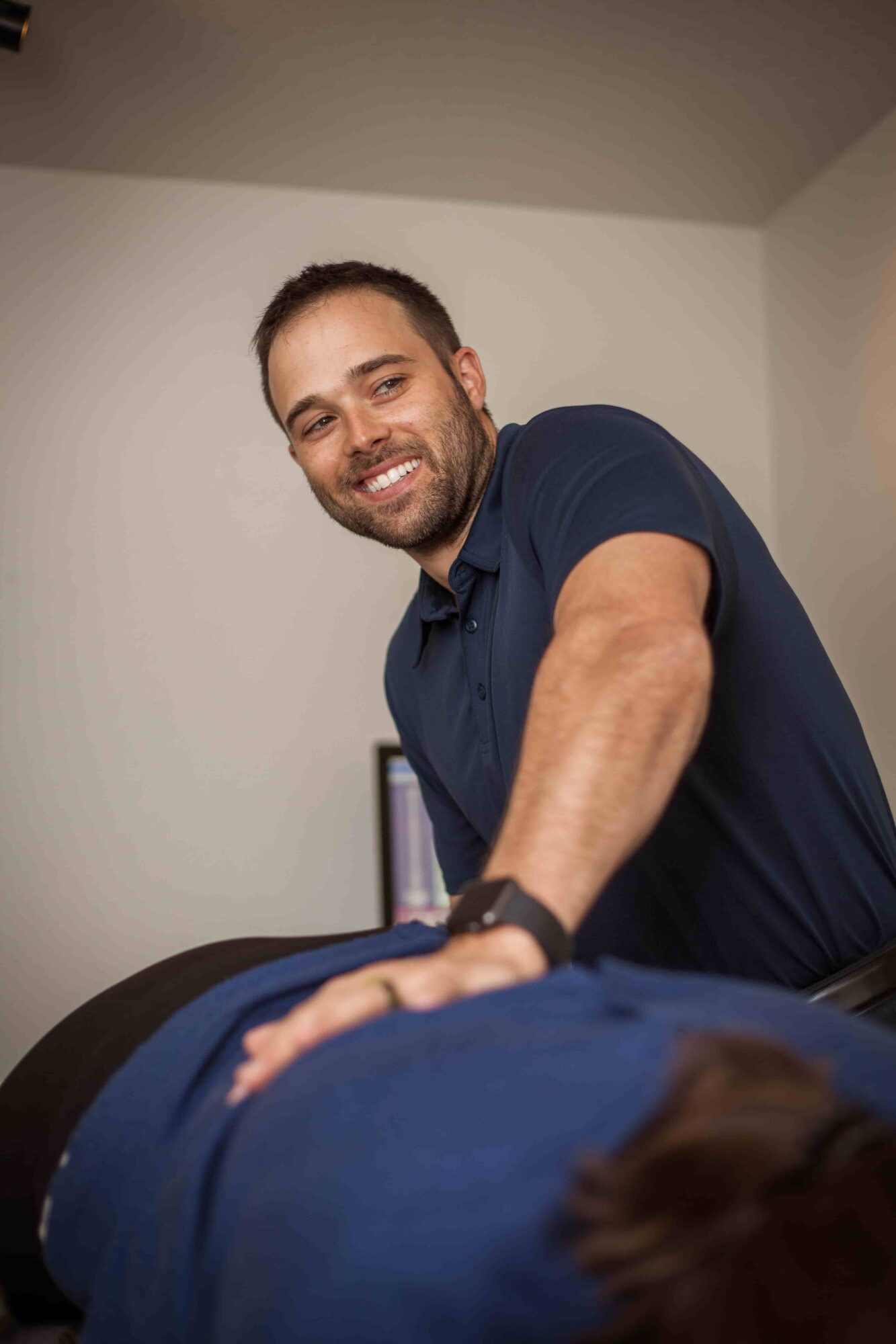 Smiling man with beard leaning over a person lying down, in a room with plain walls.