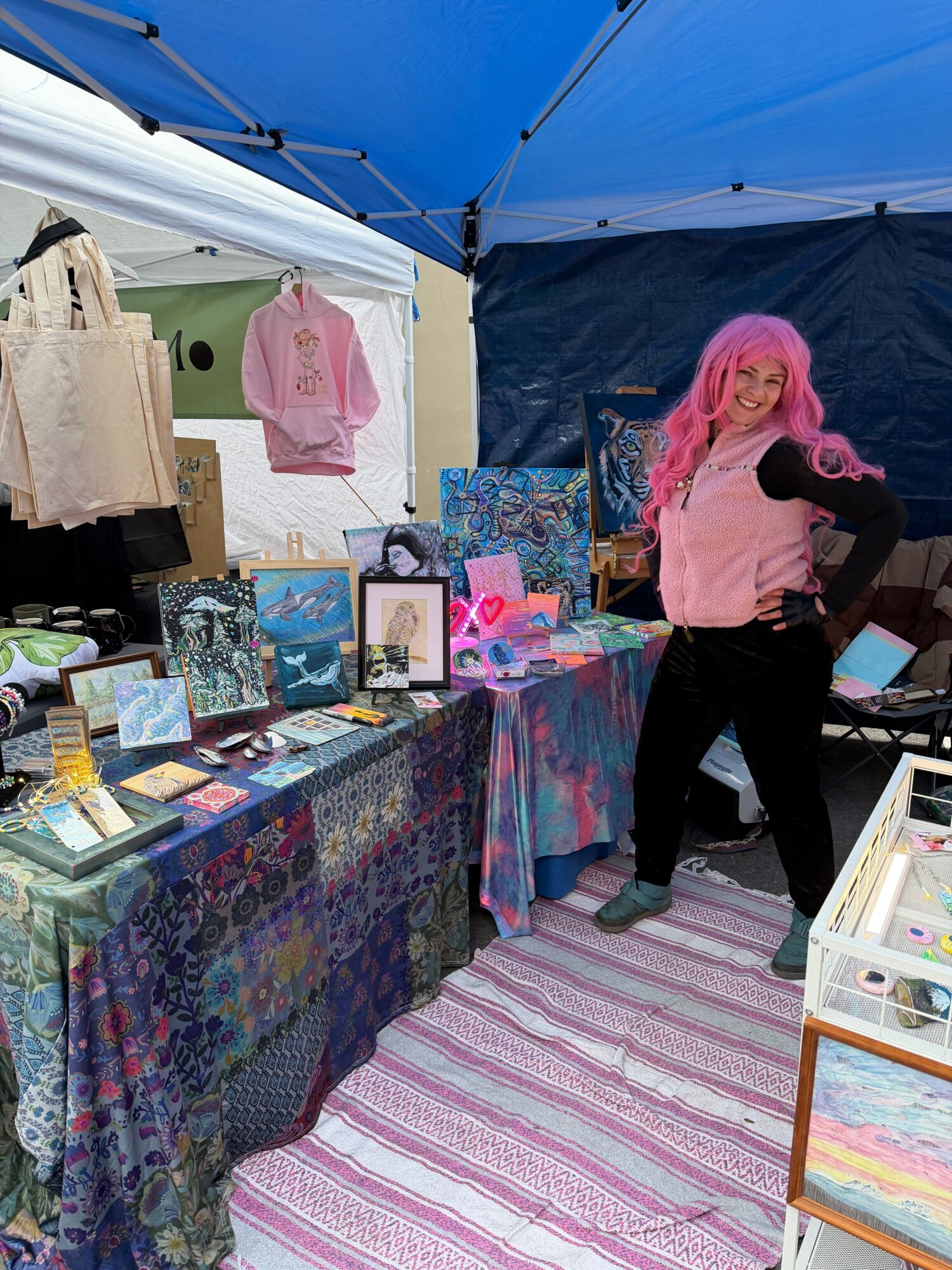Person with pink hair and pink top browsing colorful artwork at an outdoor market stall.