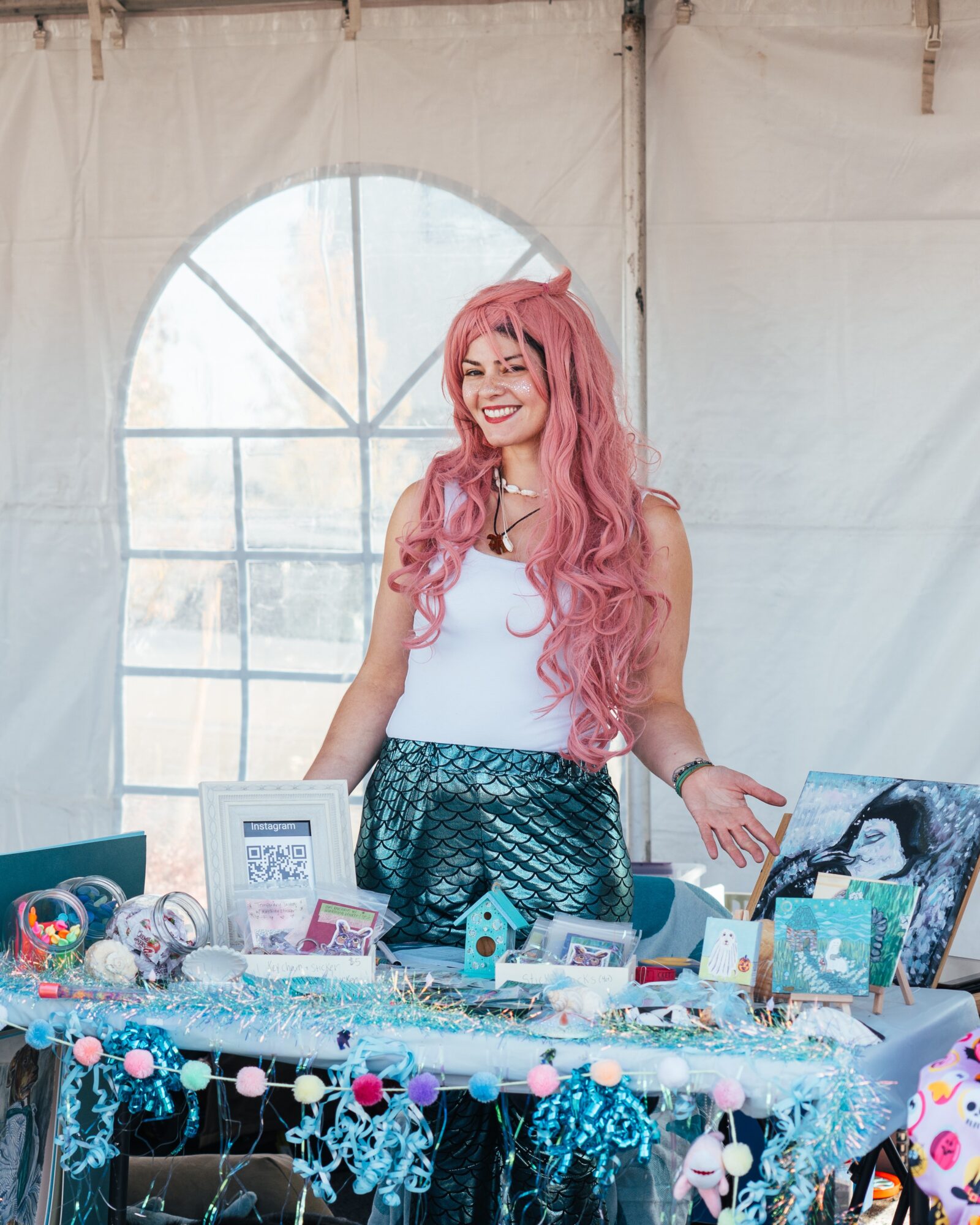 Young woman with long pink hair smiling behind a table with colorful crafts and decorations inside a tent.