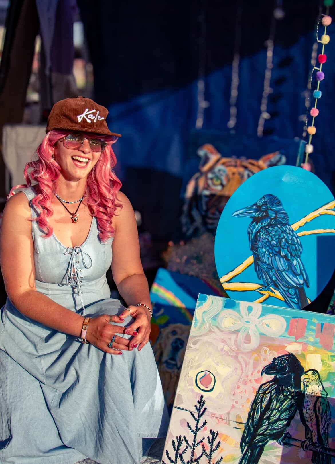 Smiling woman with pink hair, wearing a brown cap and light dress, standing near colorful artwork and decorations.