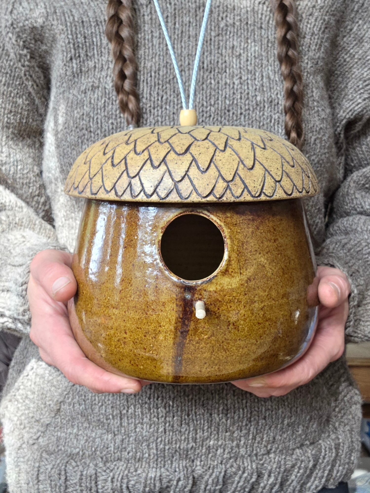 Person holding a ceramic birdhouse with a round opening, a textured roof, and a string for hanging, against a gray sweater.