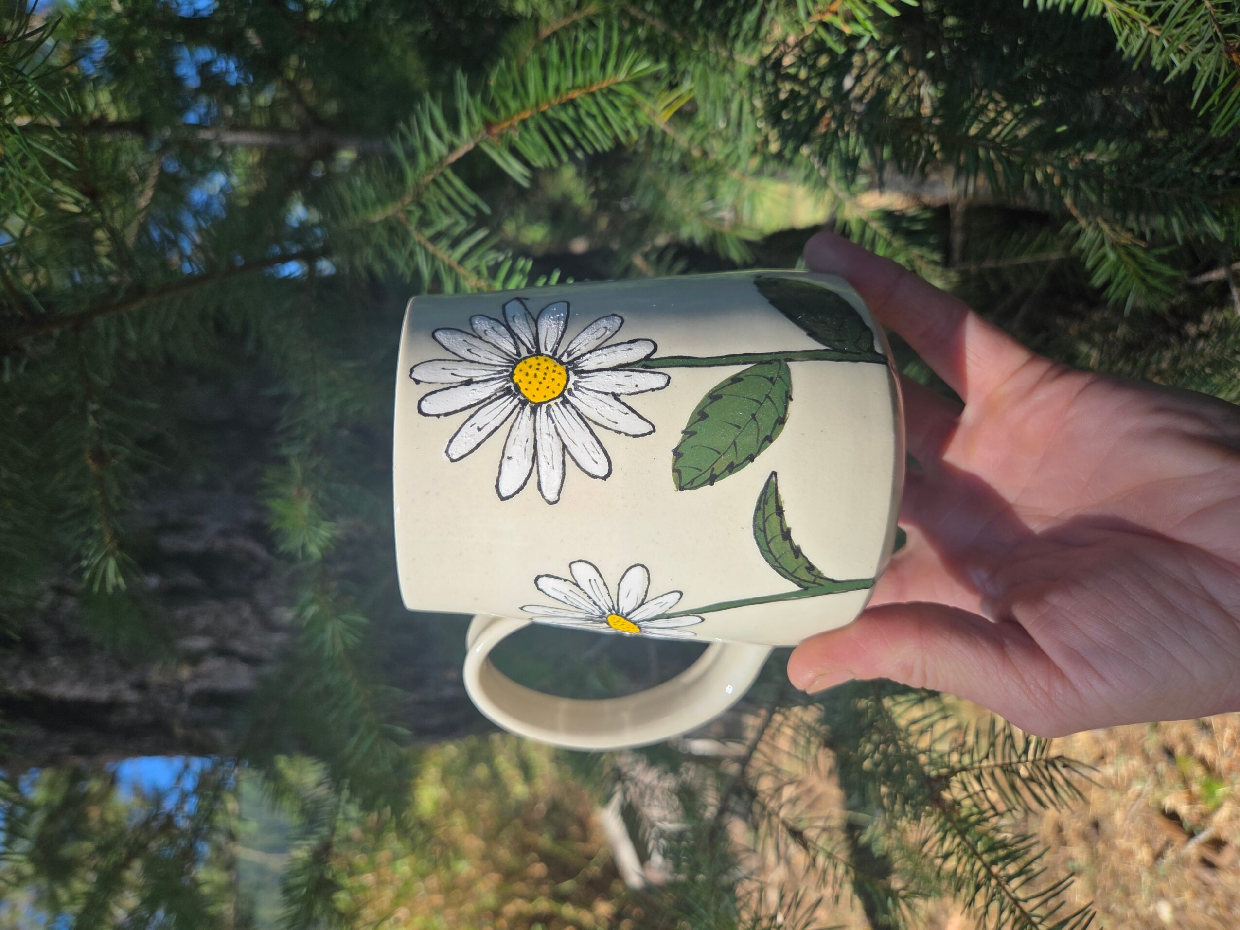 Hand holding a white mug with daisy flower and leaf design, outdoors with trees and sunlight.