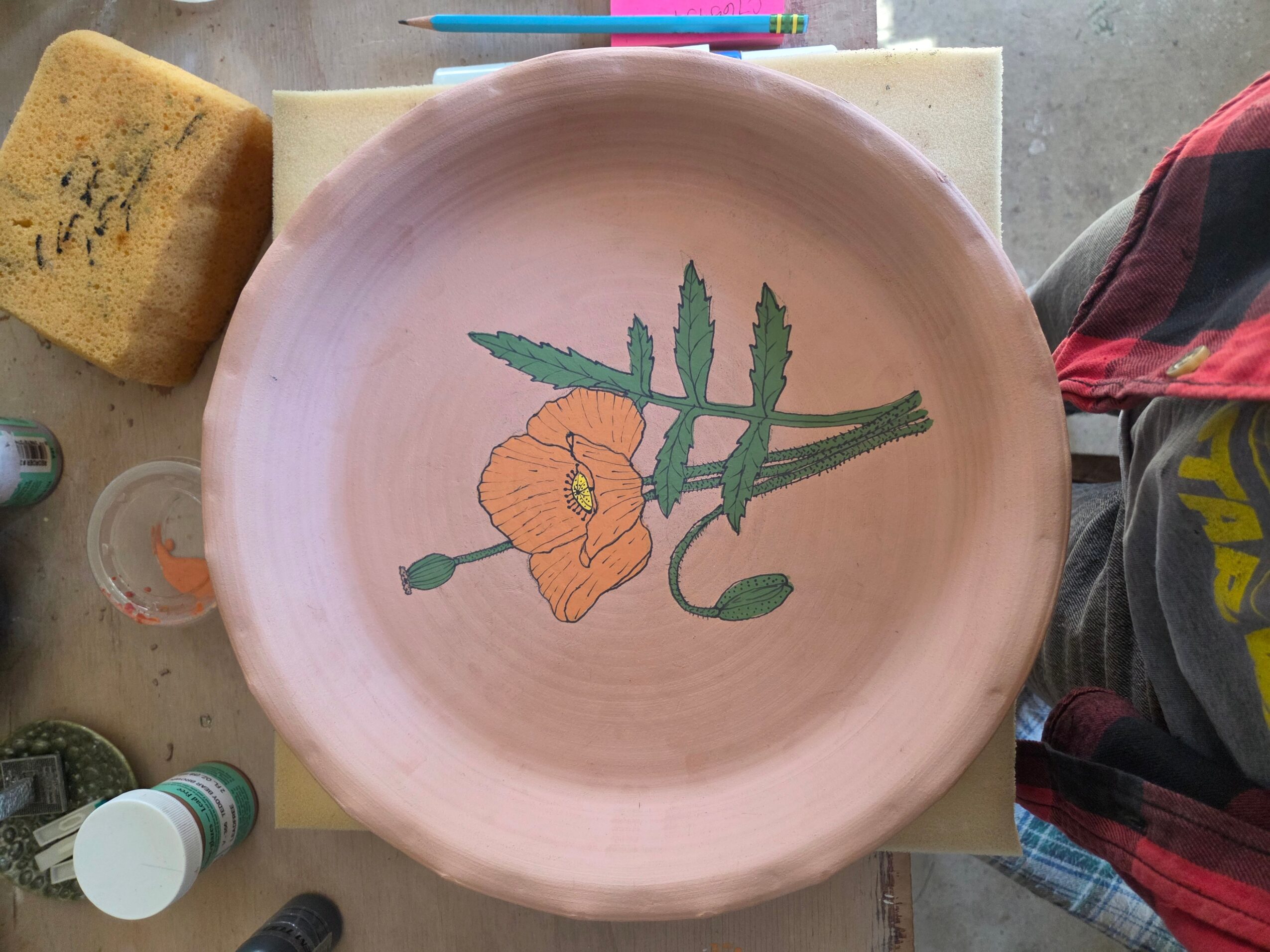 Round pink ceramic plate with a painted orange flower and green leaves, placed on a table with art supplies.