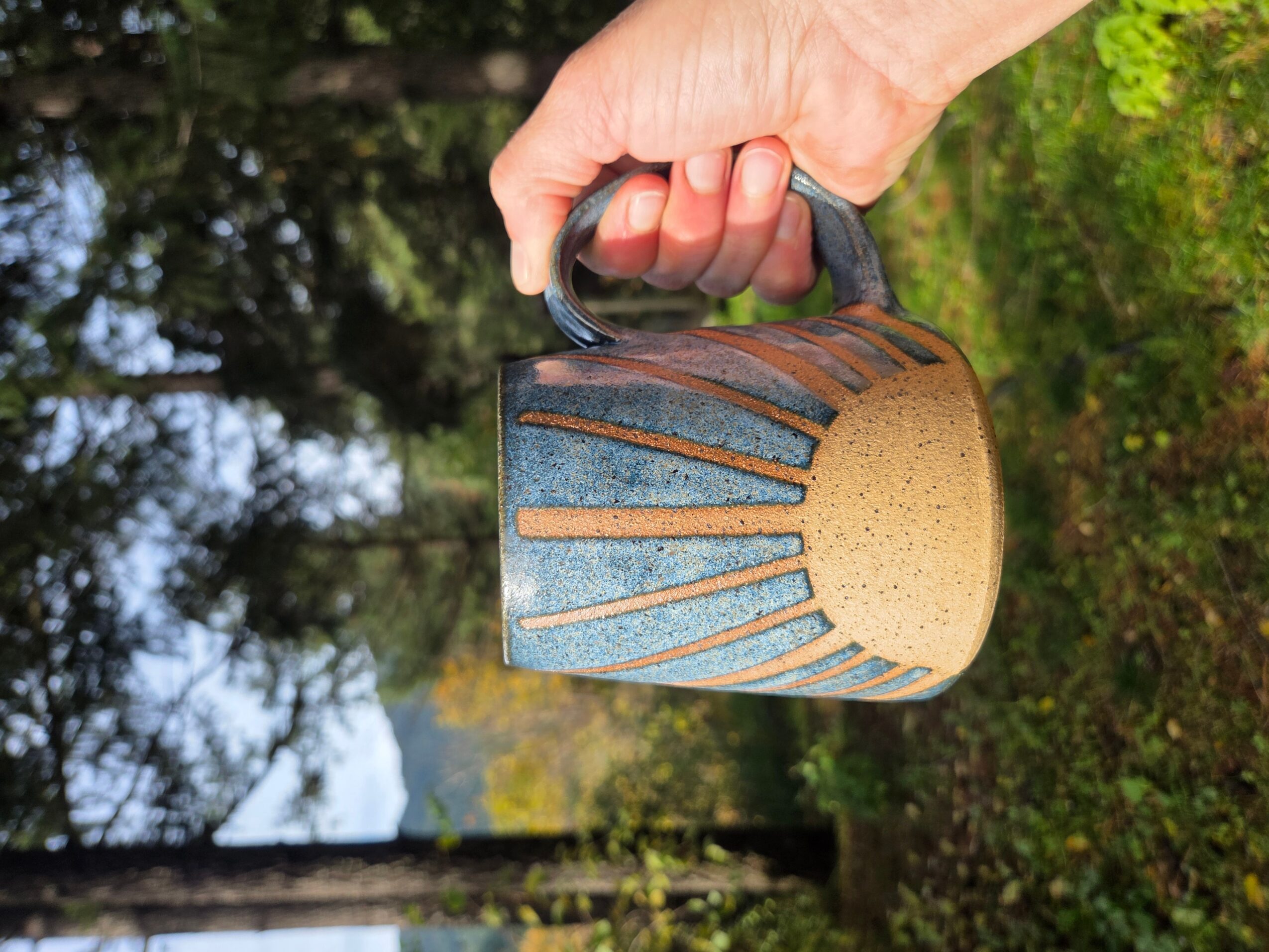 Hand holding a ceramic mug with blue and brown design, outdoors with trees and greenery in background.