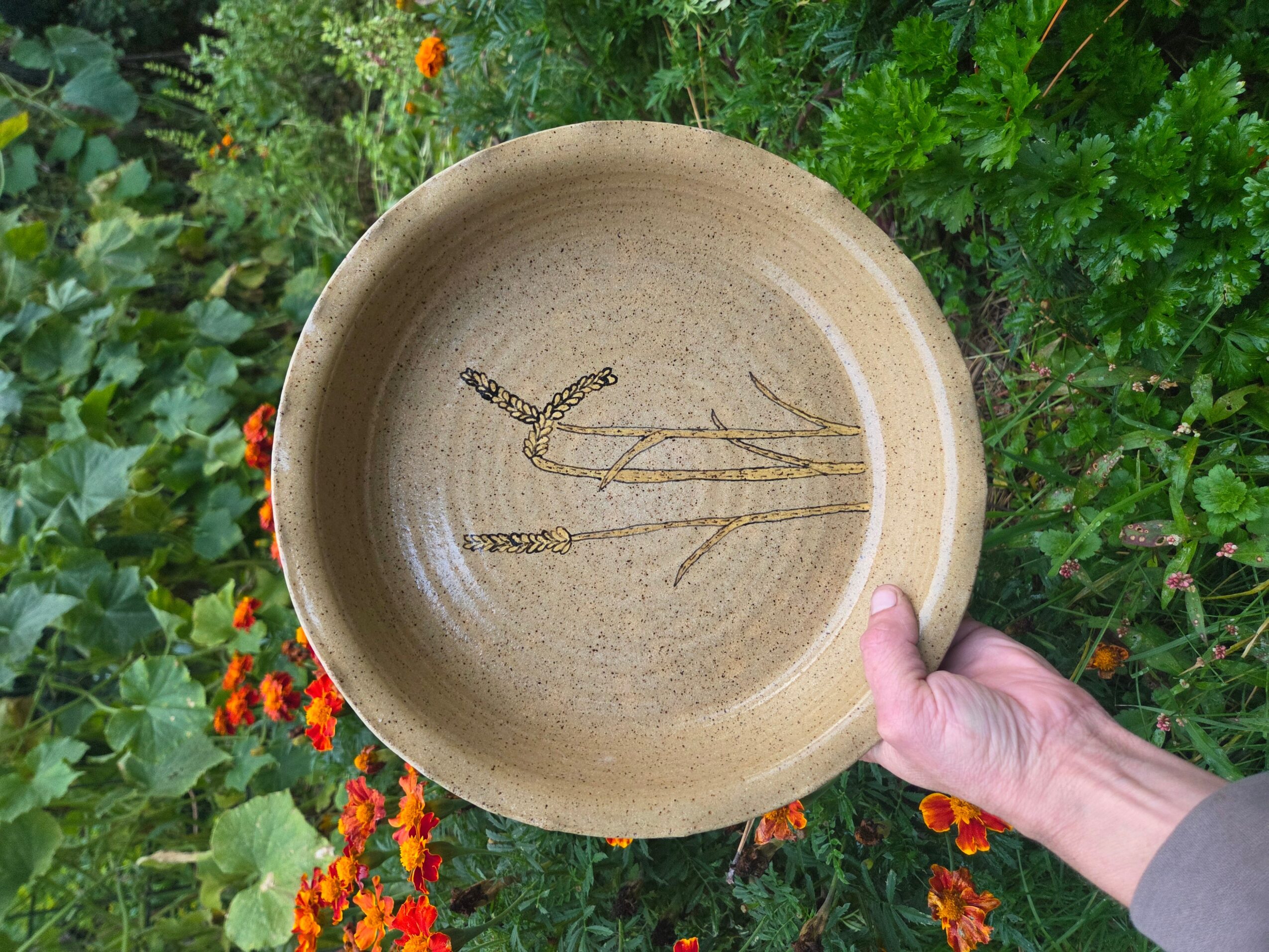 Person holding a beige ceramic bowl with a plant drawing inside, outdoors among green foliage and orange flowers.