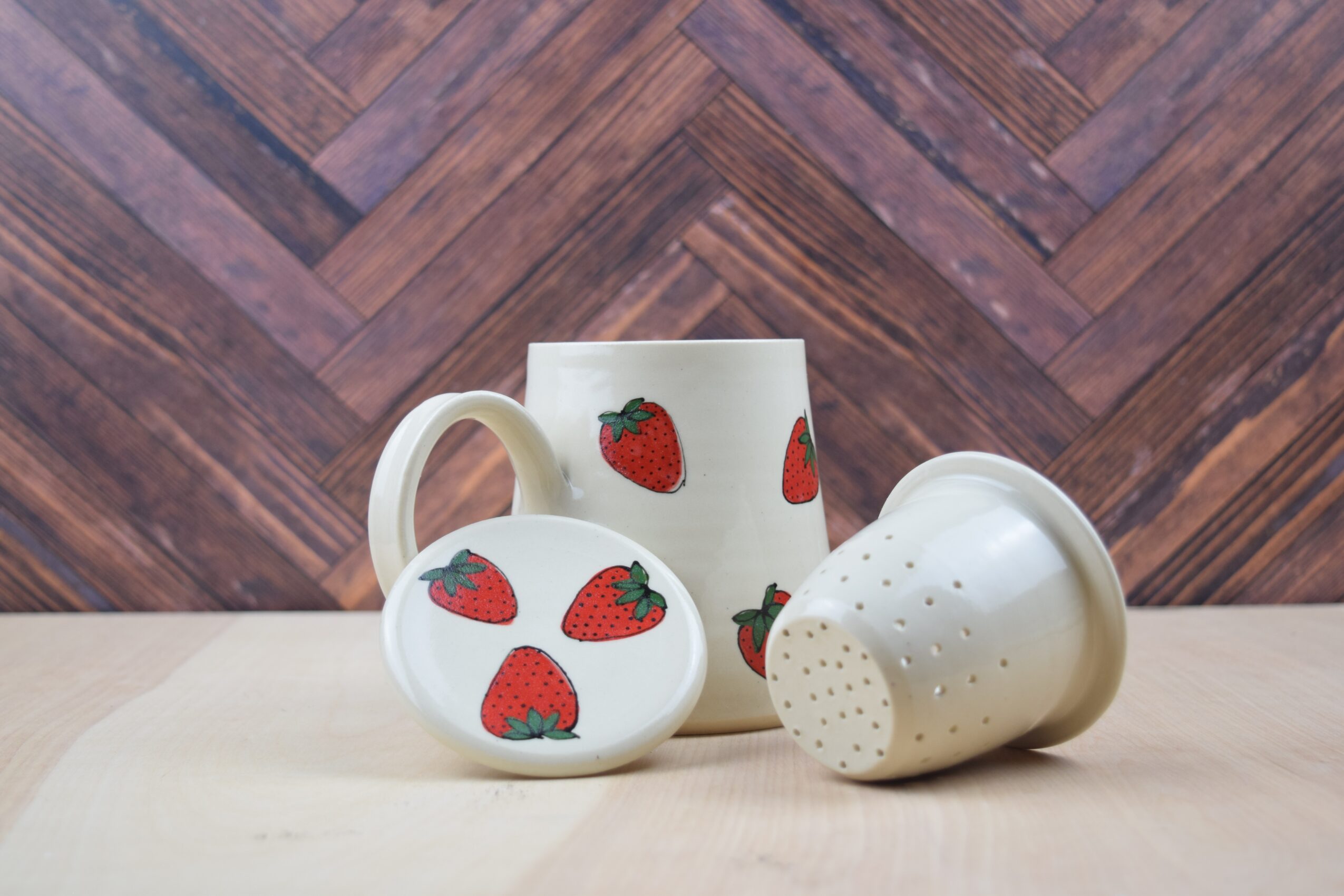 Three white ceramic items with strawberry patterns on a light surface, with a wooden patterned background.
