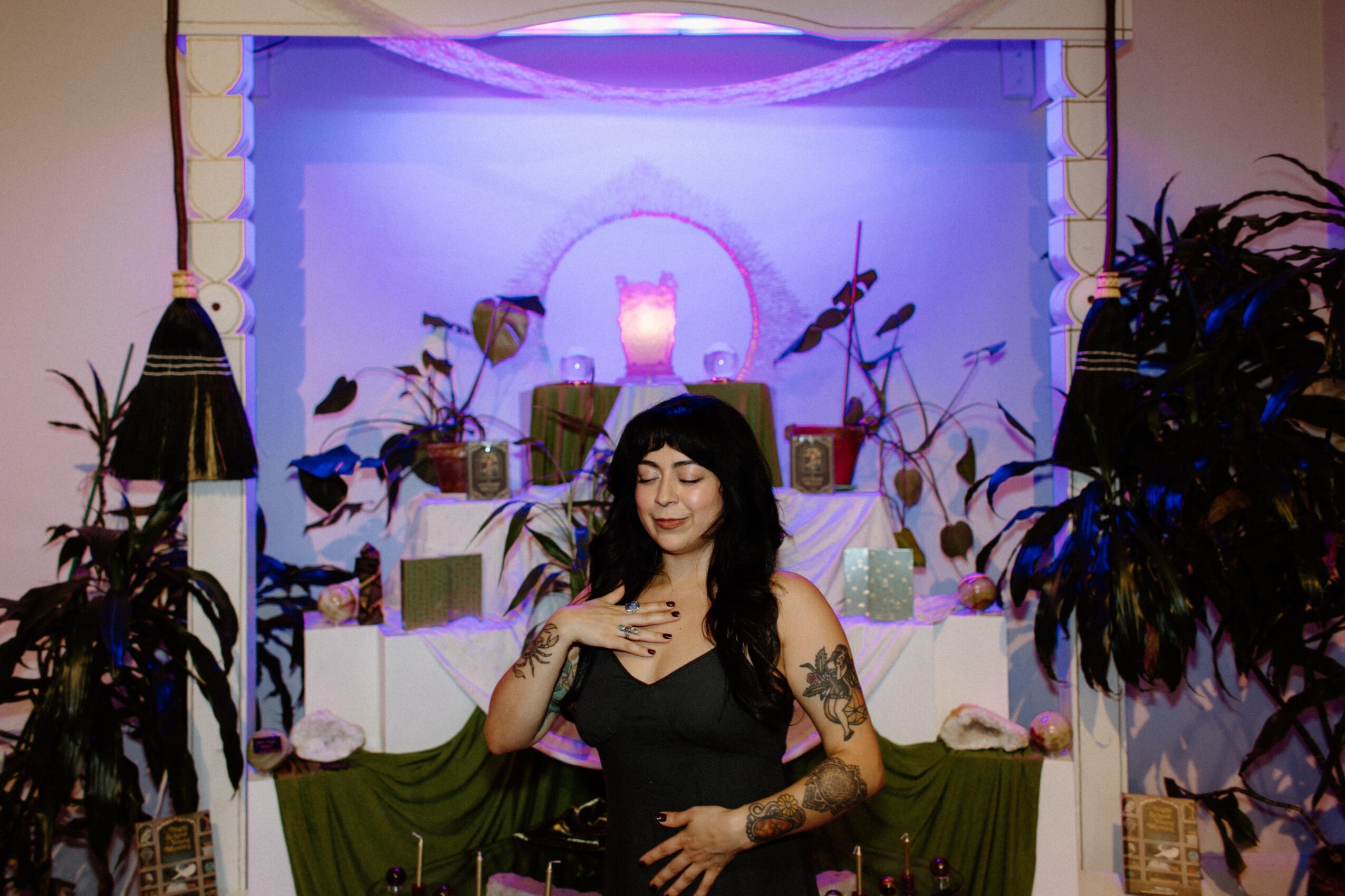 Woman with dark hair and tattoos in a black dress stands in front of a decorated backdrop with plants and candles.