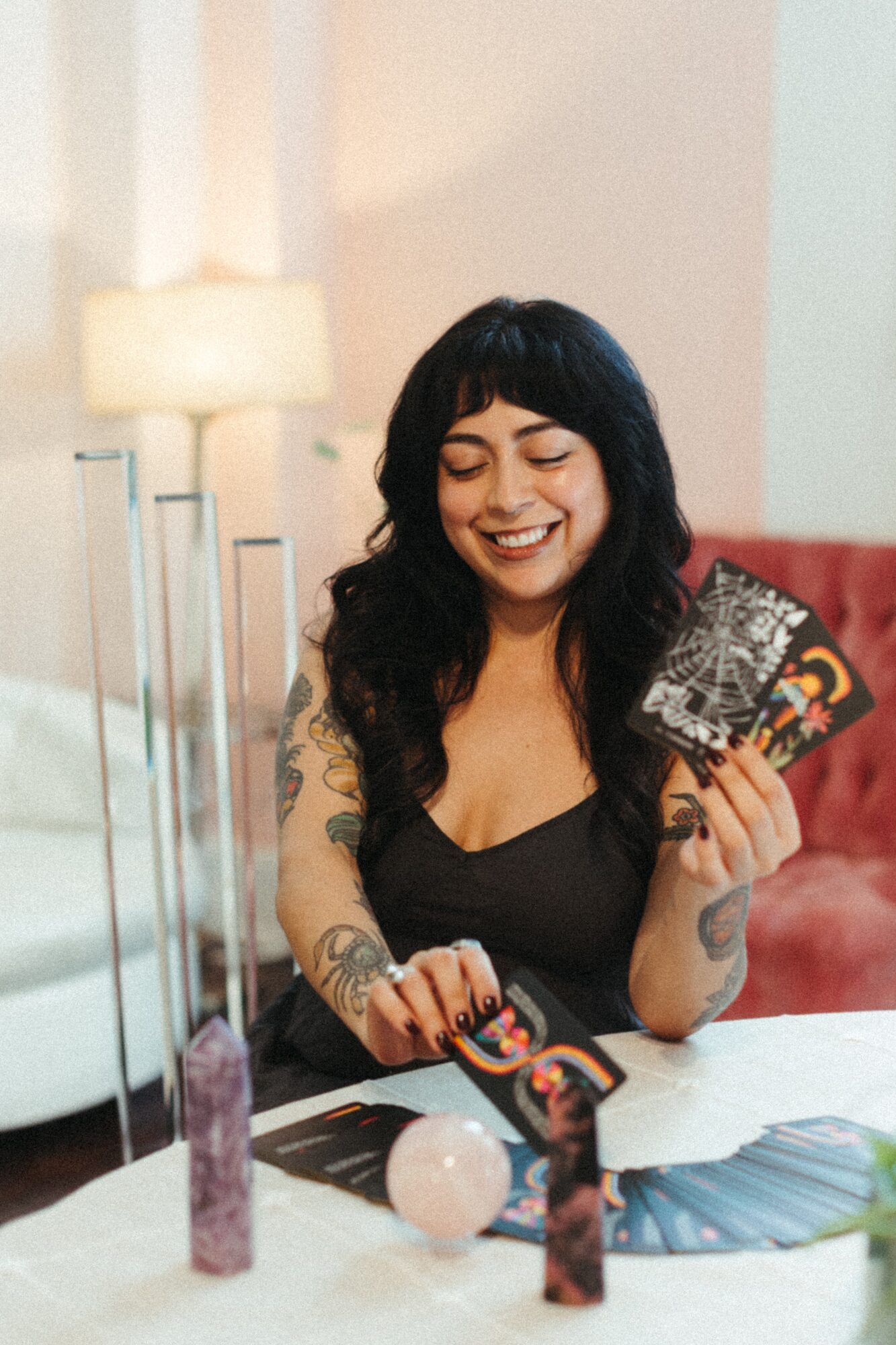 Smiling woman with black hair and tattoos holding a card, sitting at a table with various objects, in a cozy room.