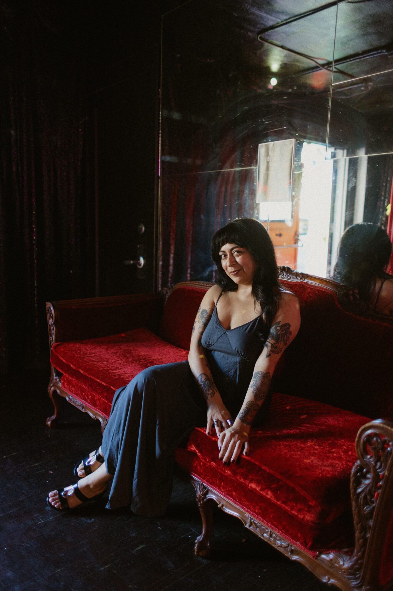 Woman sitting on a red velvet sofa in a dimly lit room with mirrors, sunlight coming through a window behind her.