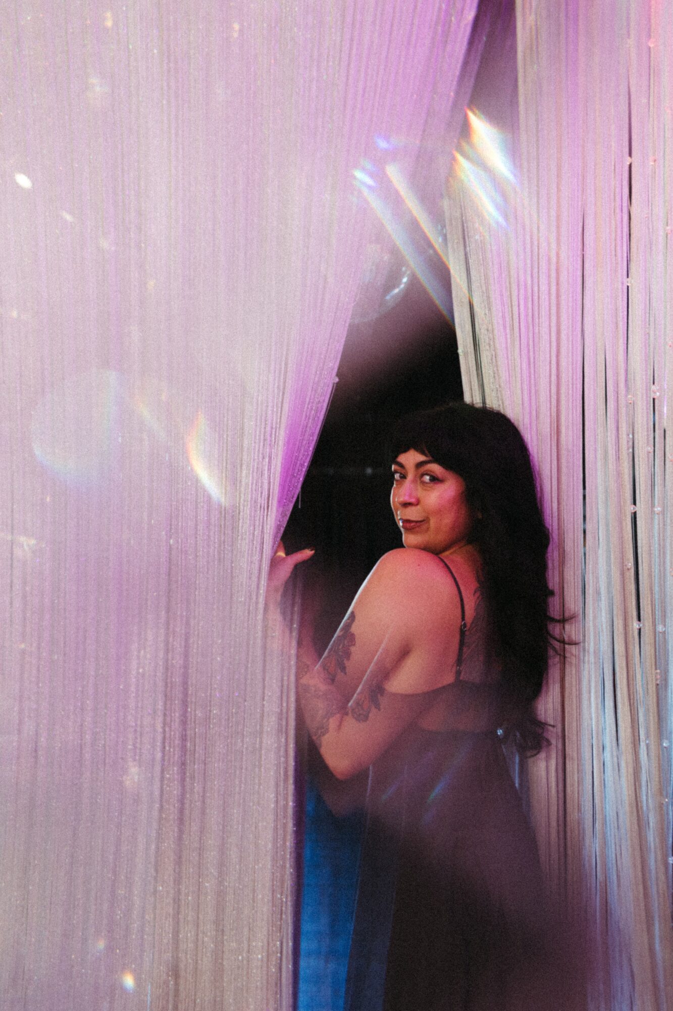 Woman with dark hair and a sleeveless top stands between shimmering curtains, looking over her shoulder at the camera.