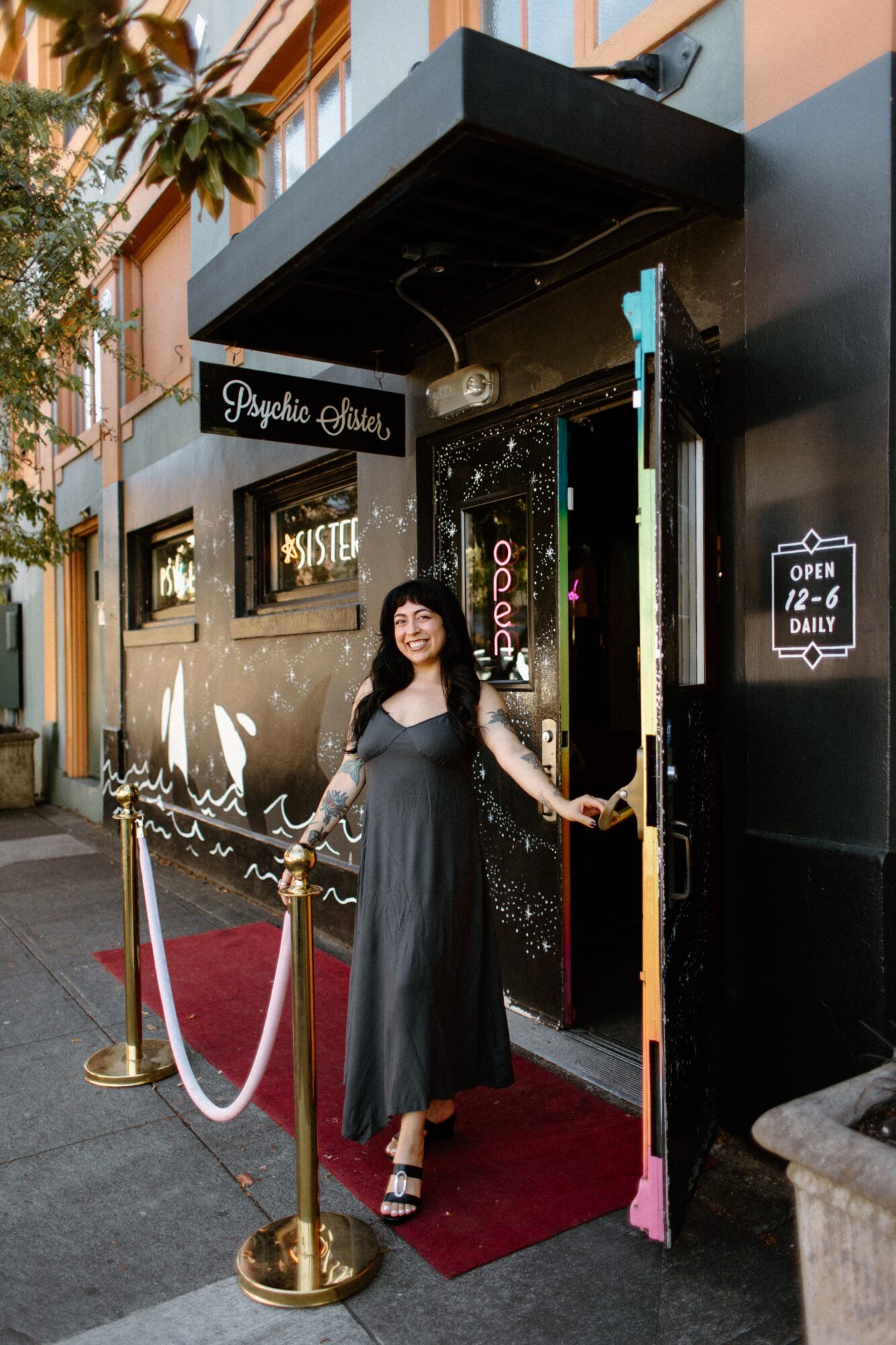 Woman in black dress standing on red carpet outside a building entrance with a sign, ropes, and a colorful door.