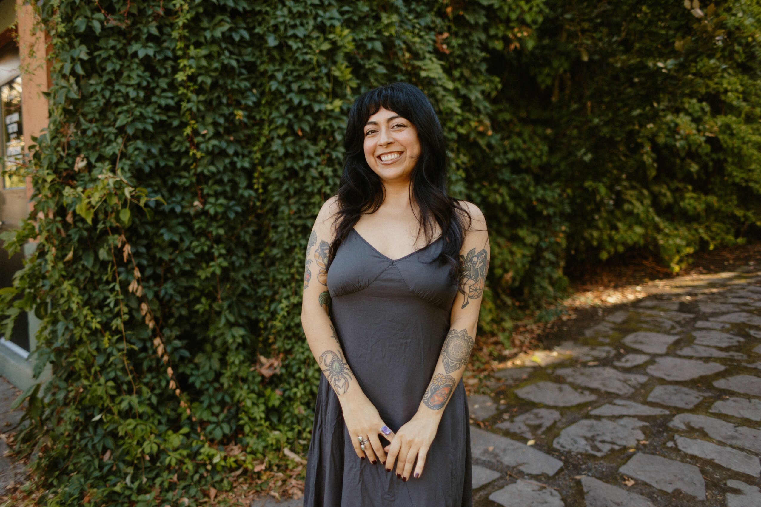 Smiling woman with dark hair and tattoos in a gray dress outdoors, surrounded by greenery and stone path.