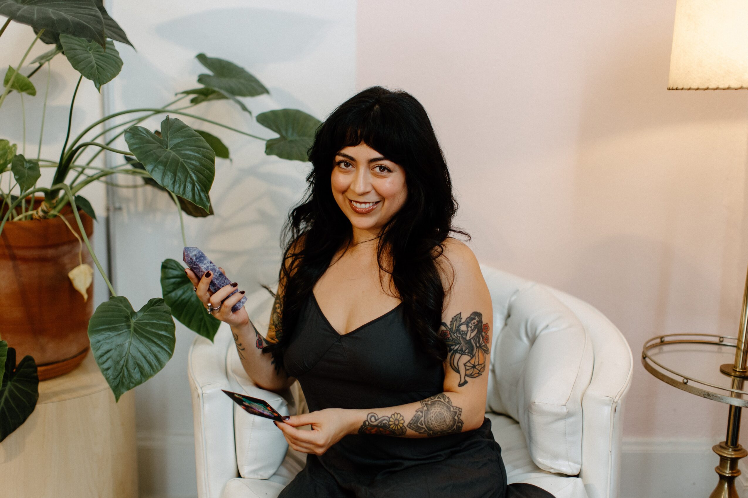 Woman with dark hair and tattoos smiling, holding a phone, sitting on a white armchair, surrounded by plants and a lamp.
