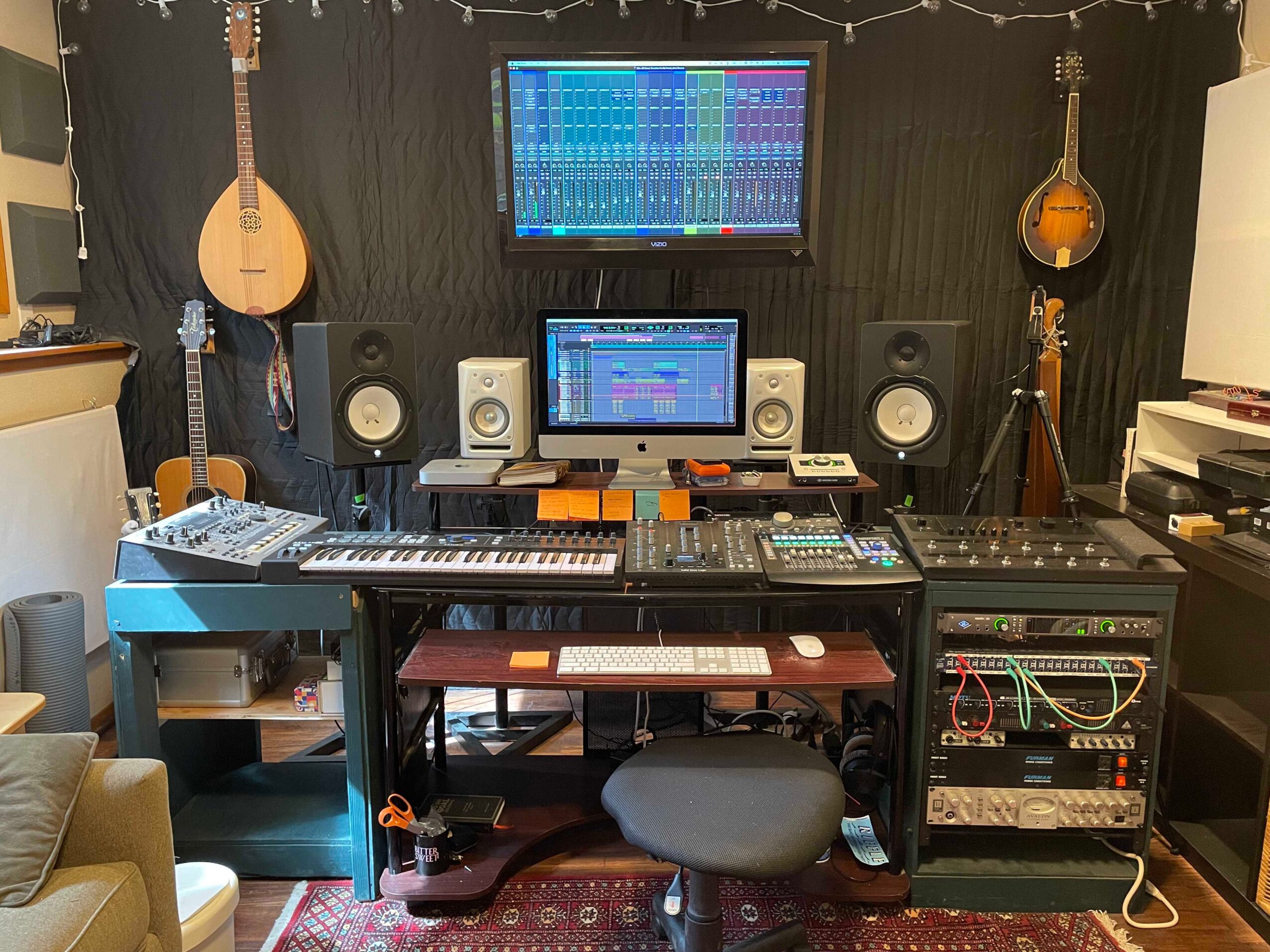 Music studio with computer, keyboard, speakers, guitars, and audio equipment on black wall, with monitors displaying digital audio workstations.