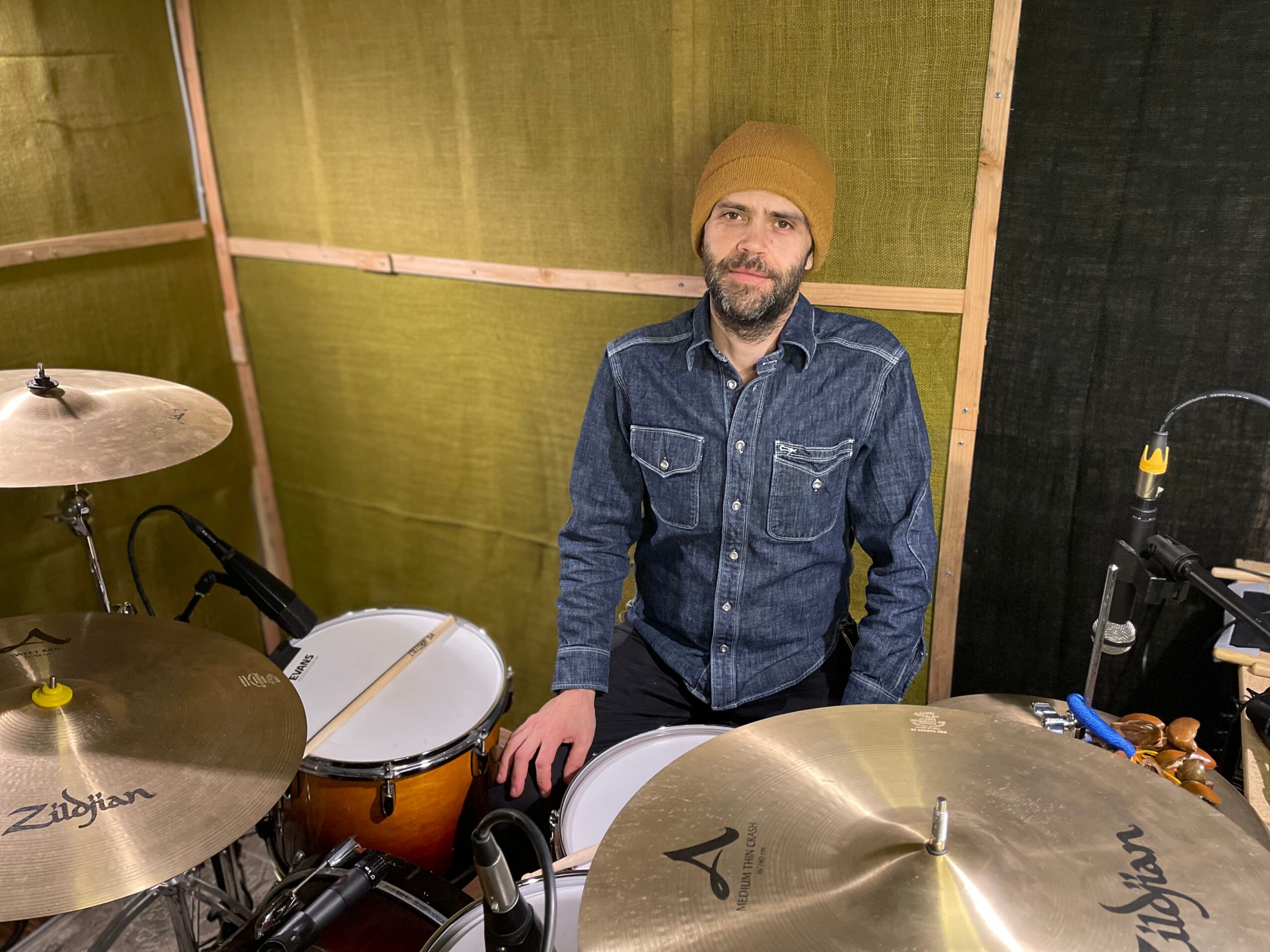 Man with a beard wearing a beige beanie and denim jacket, standing behind drums in a music studio.