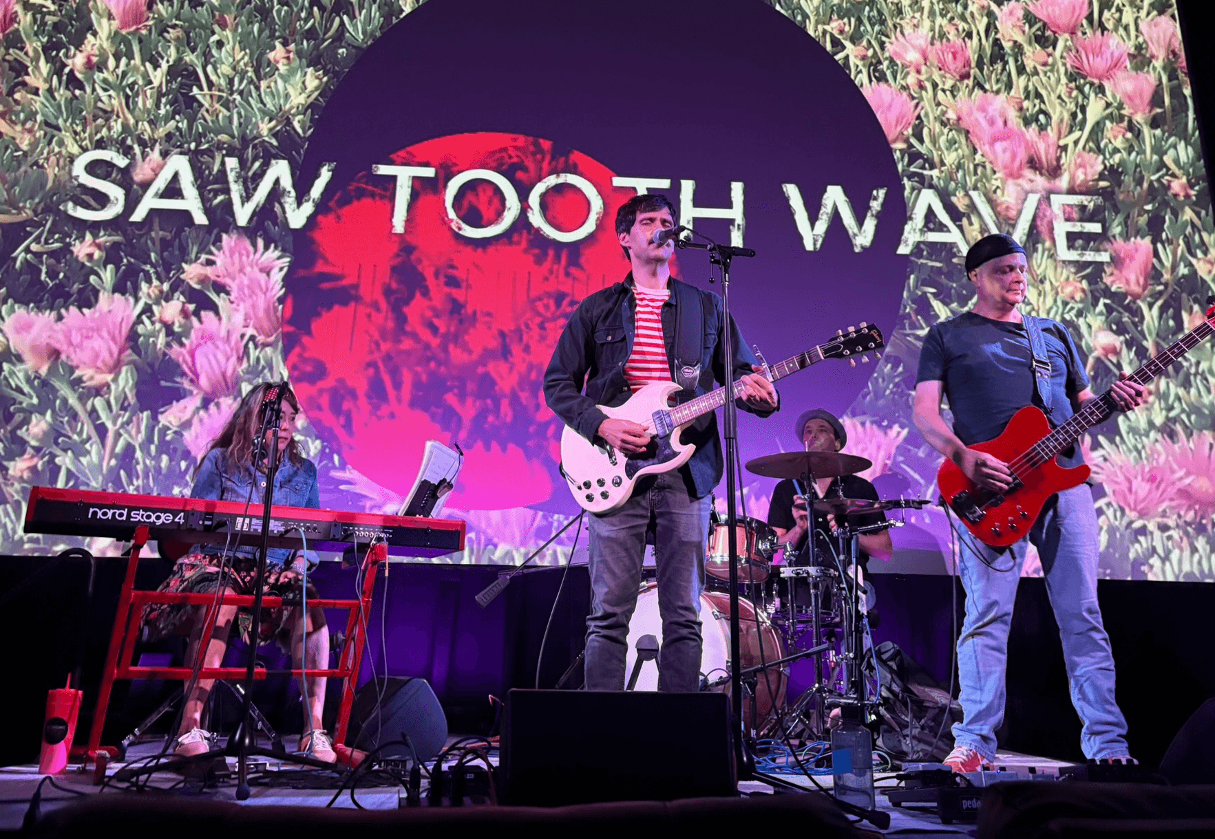Band performing on stage with a large screen behind displaying flowers and text, four musicians playing instruments.
