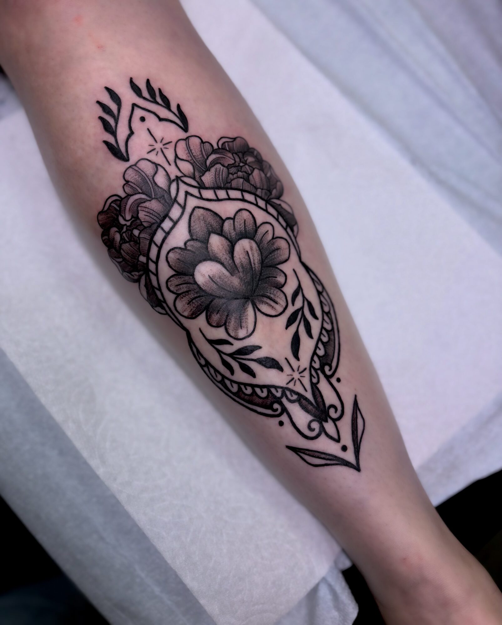 Black floral tattoo on forearm with detailed flowers and leaves, enclosed in a decorative frame, on light skin.
