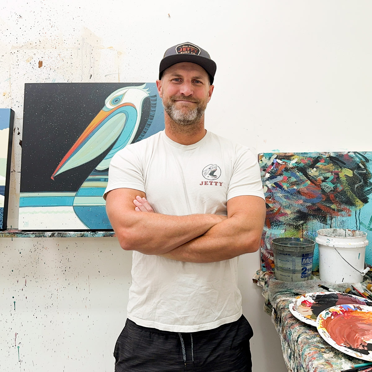 Man with a beard wearing a white t-shirt and a cap, standing with arms crossed in front of paintings and art supplies.