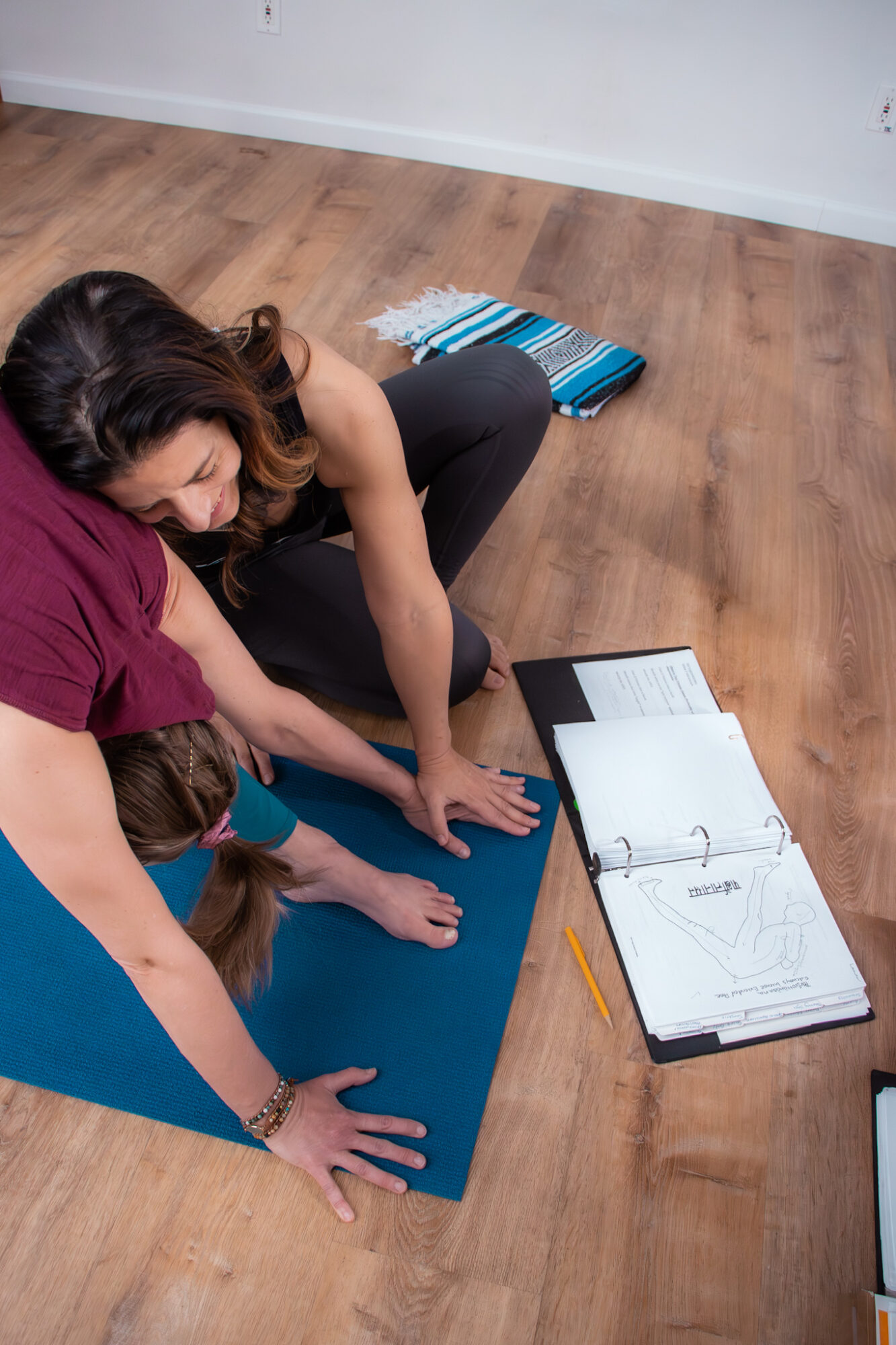 Two women practicing yoga on a blue mat indoors, with notebooks and a pencil nearby.