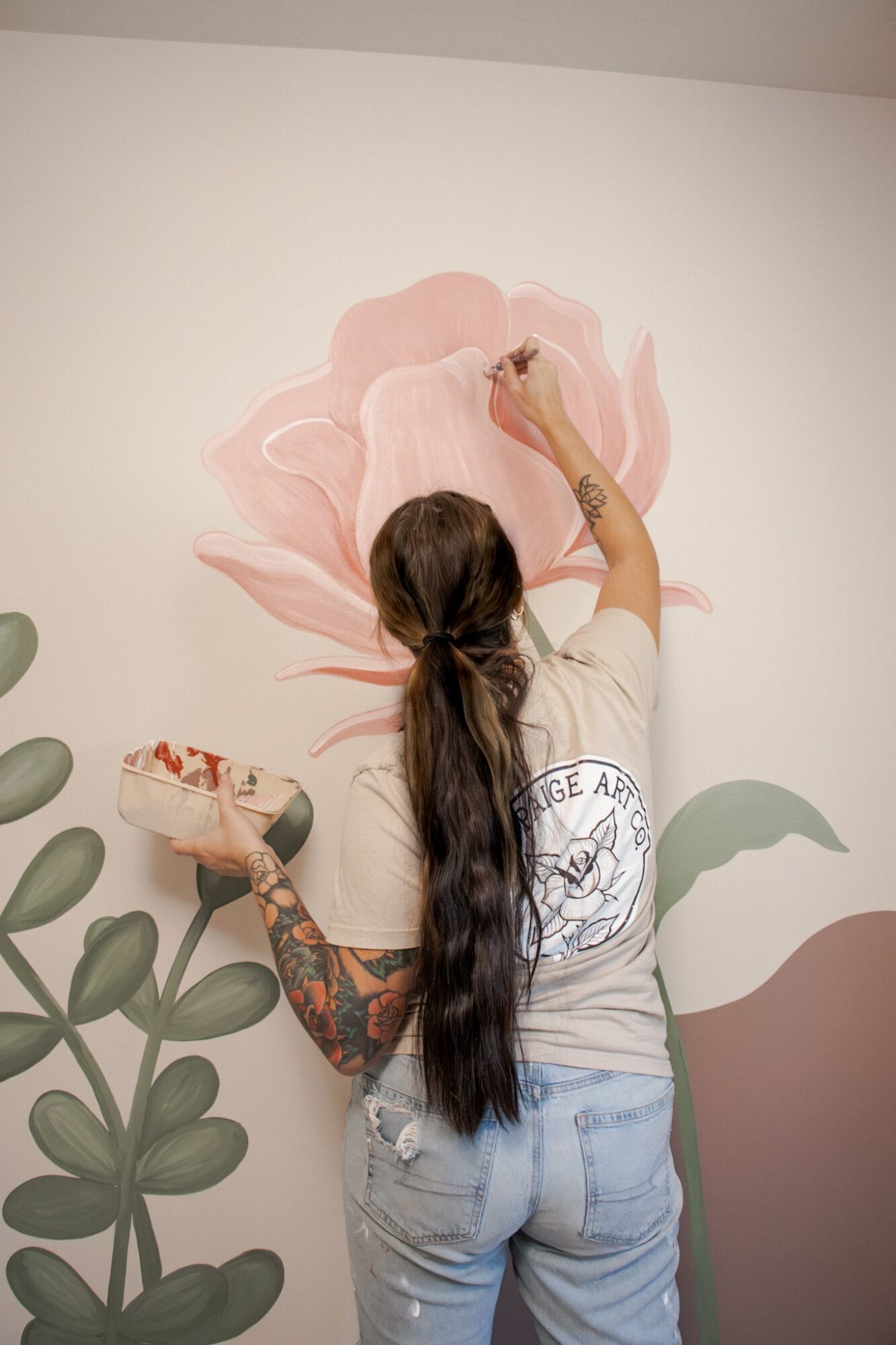 Person with long dark hair and tattoos painting a large pink flower on a wall, holding a paintbrush and palette.
