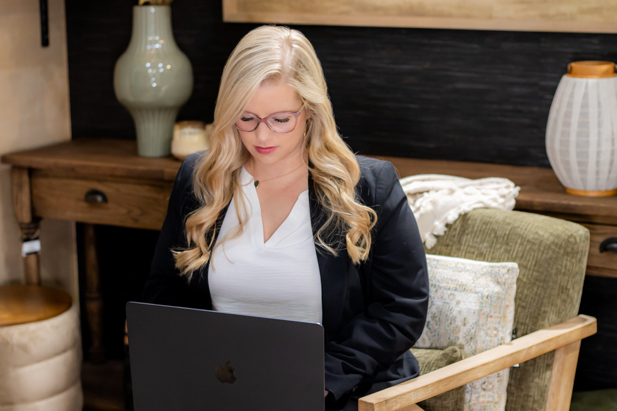Woman with long blonde hair and glasses working on a laptop in a cozy room with wooden furniture and decorative items.