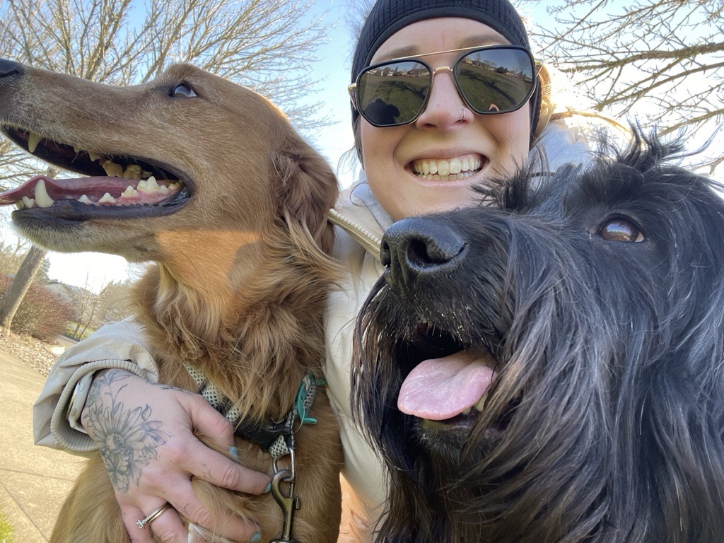 Person wearing sunglasses and a beanie smiling with two dogs outdoors on a sunny day.