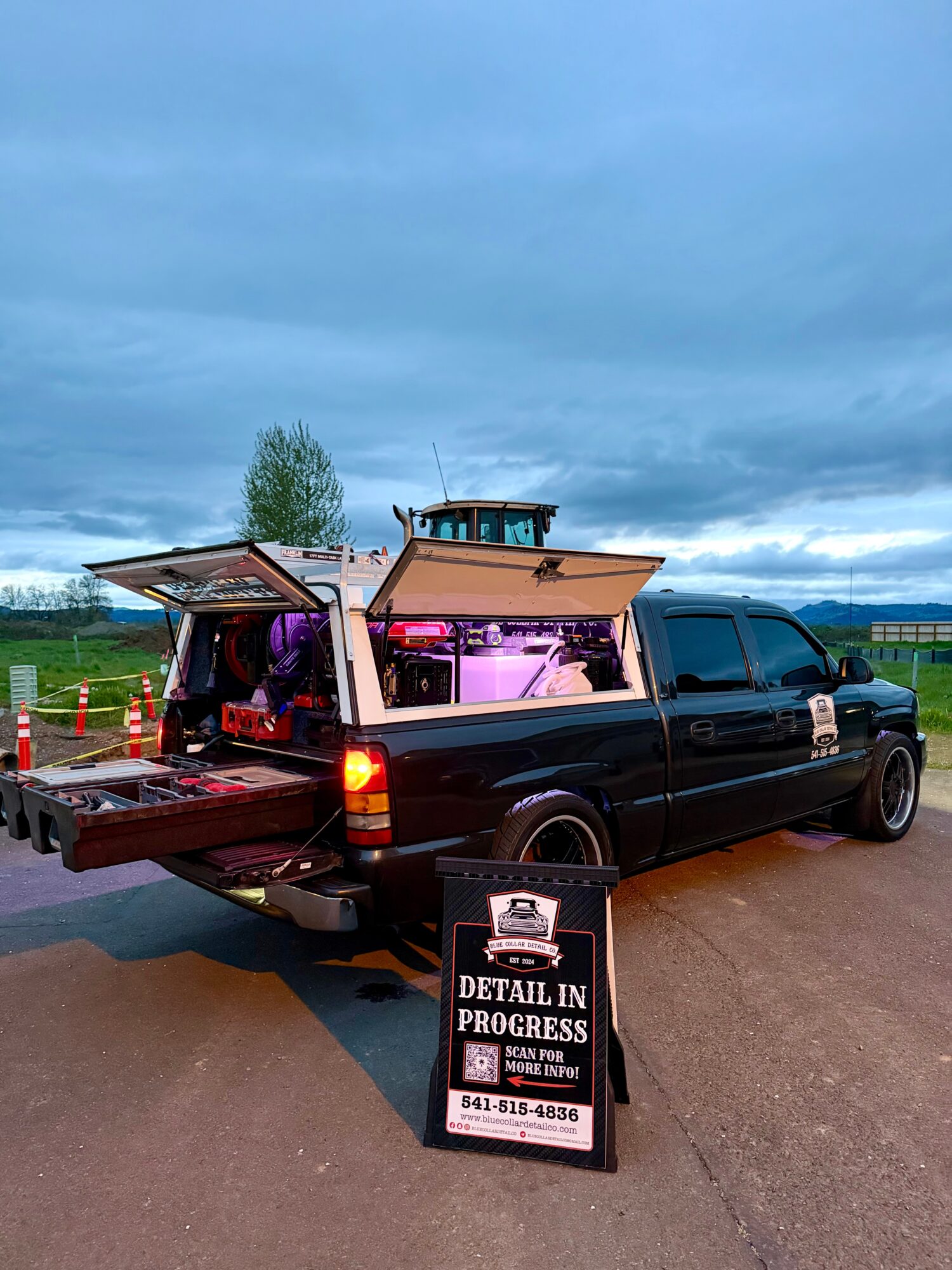 Black pickup truck with boat on trailer parked outdoors under cloudy sky, with a signboard in front advertising a detail service.