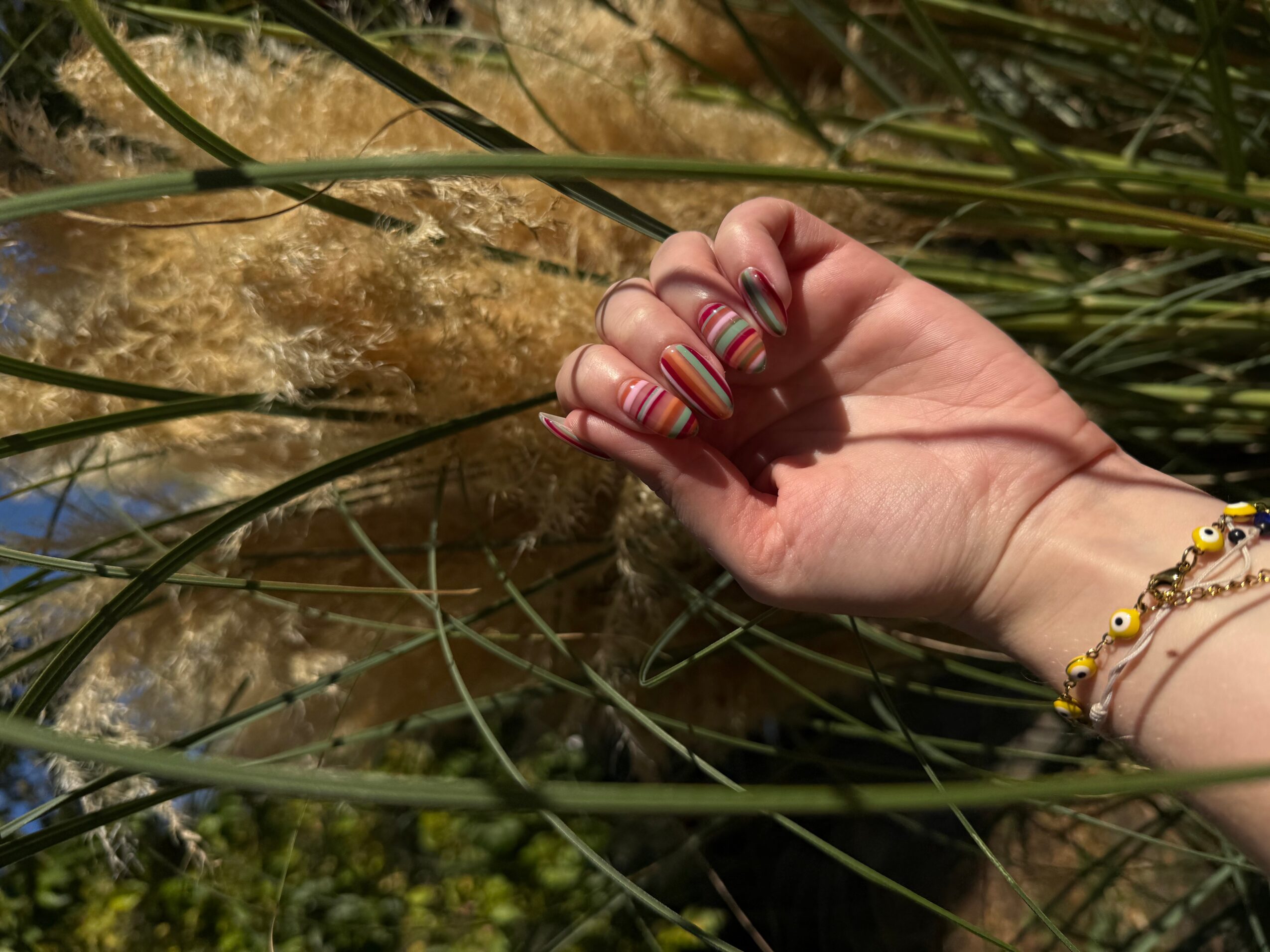 Hand with colorful striped nails near water and tall grass, wearing bracelets, outdoors.