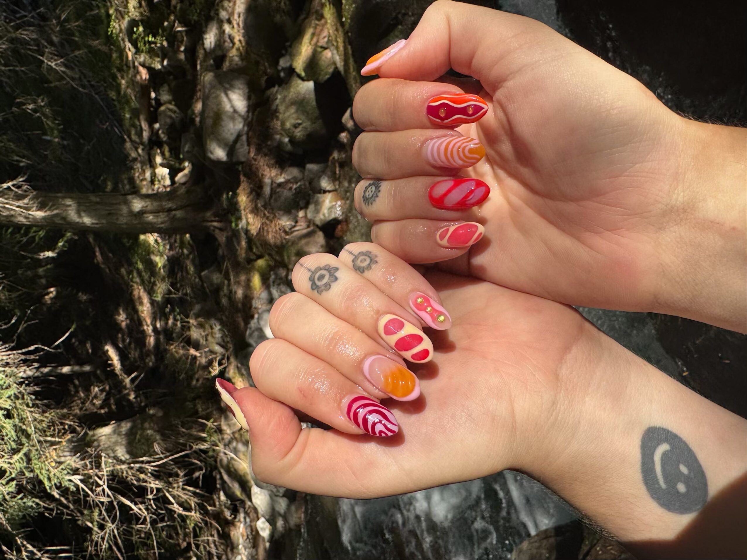 Two hands with colorful, patterned nail art, one with a tattoo of a smiley face on the wrist, outdoors near rocks and water.