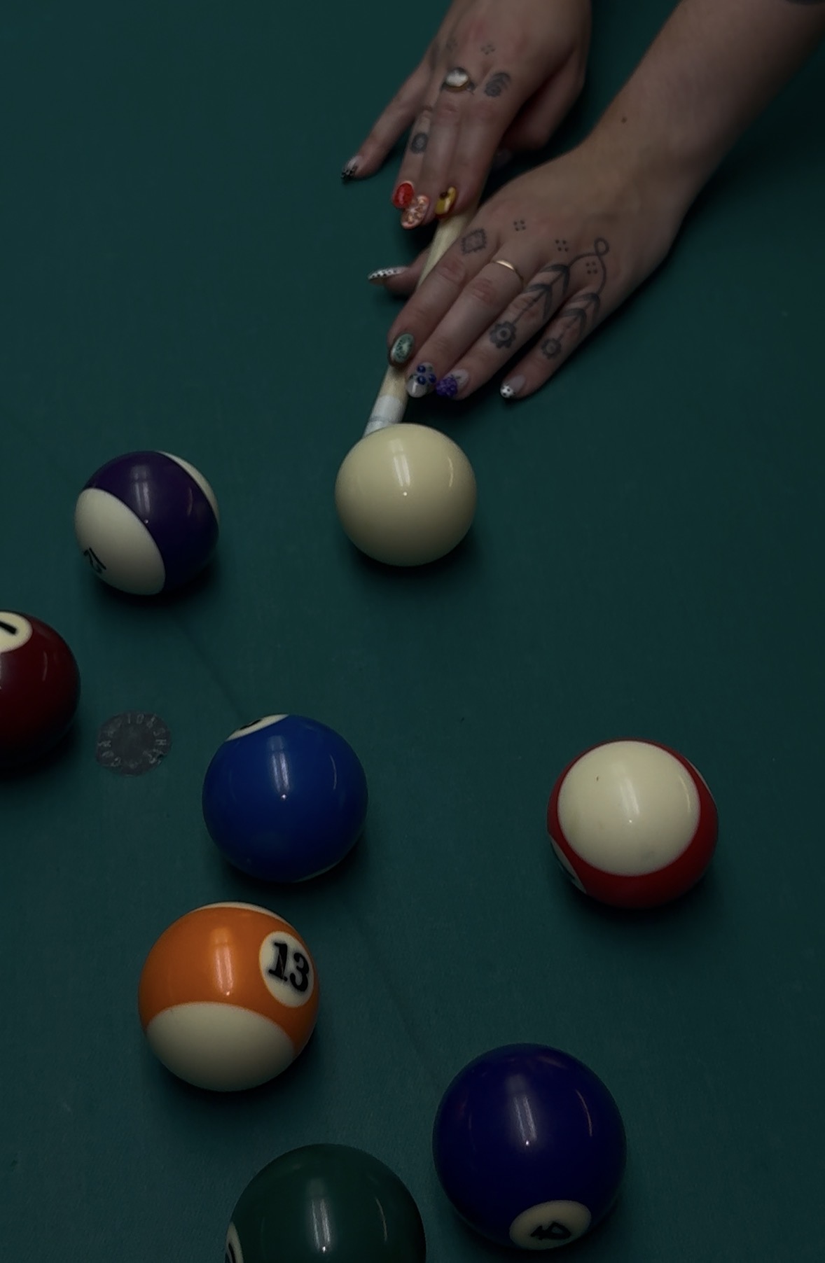 Hands with rings and painted nails reach over a pool table with billiard balls scattered, including a striped ball and solid balls.