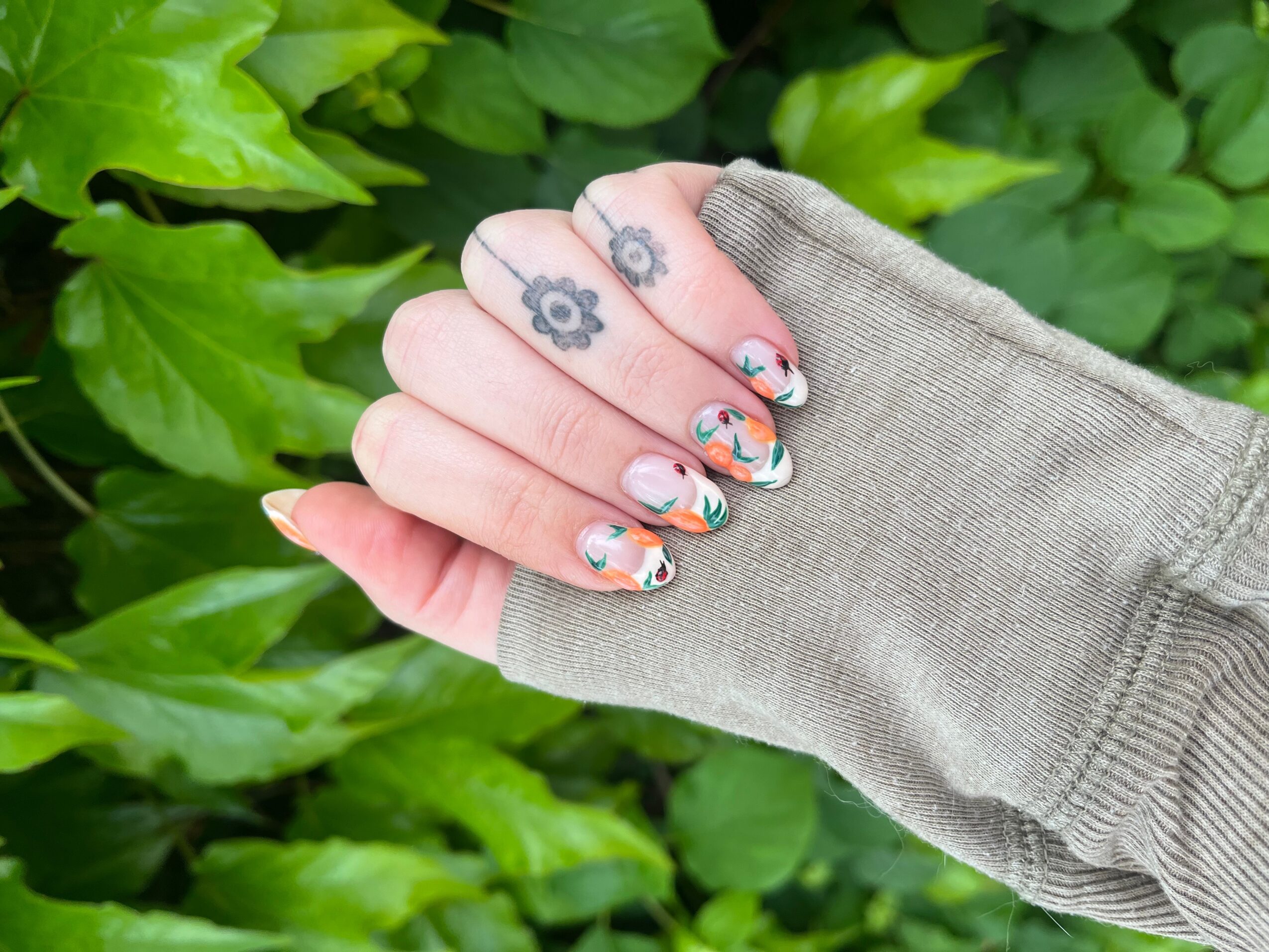 Hand with decorated nails in a gray sleeve, green leaves in background, nails have floral and colorful designs.