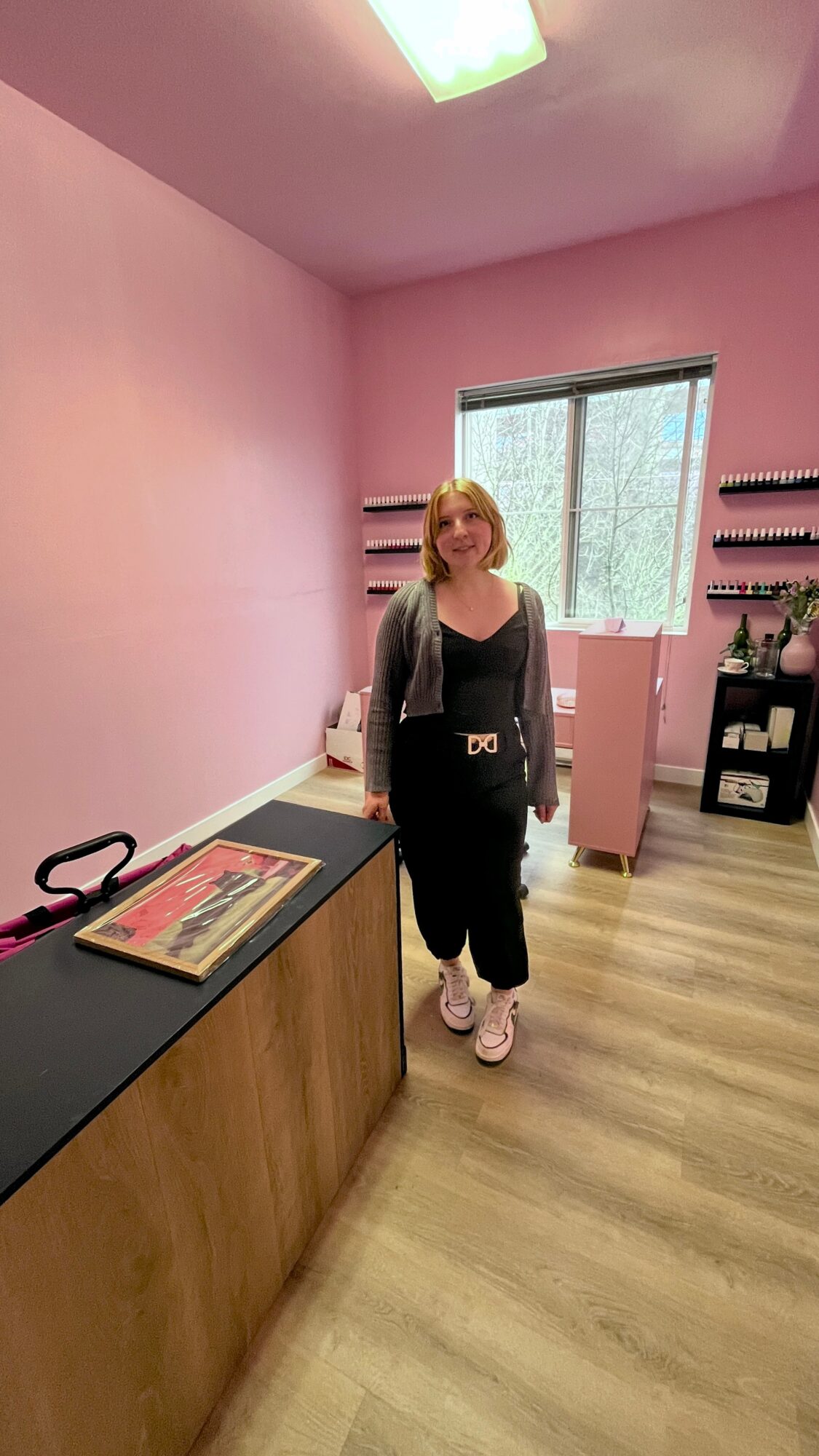 Woman with blonde hair standing in a pink room near a window, with a black and wood counter and shelves behind her.