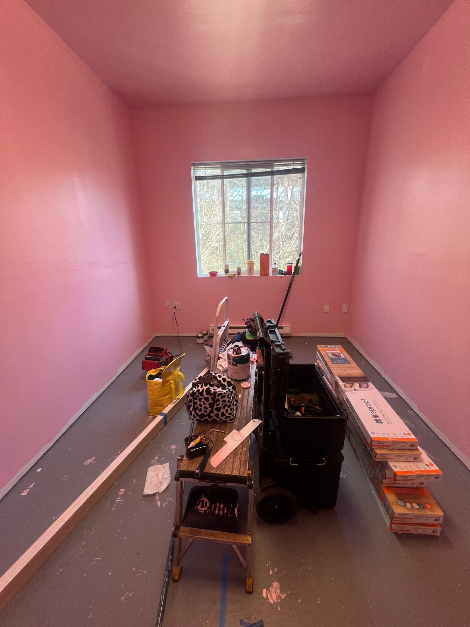 Room with pink walls, window with blinds, and various electronic equipment and boxes on a table.