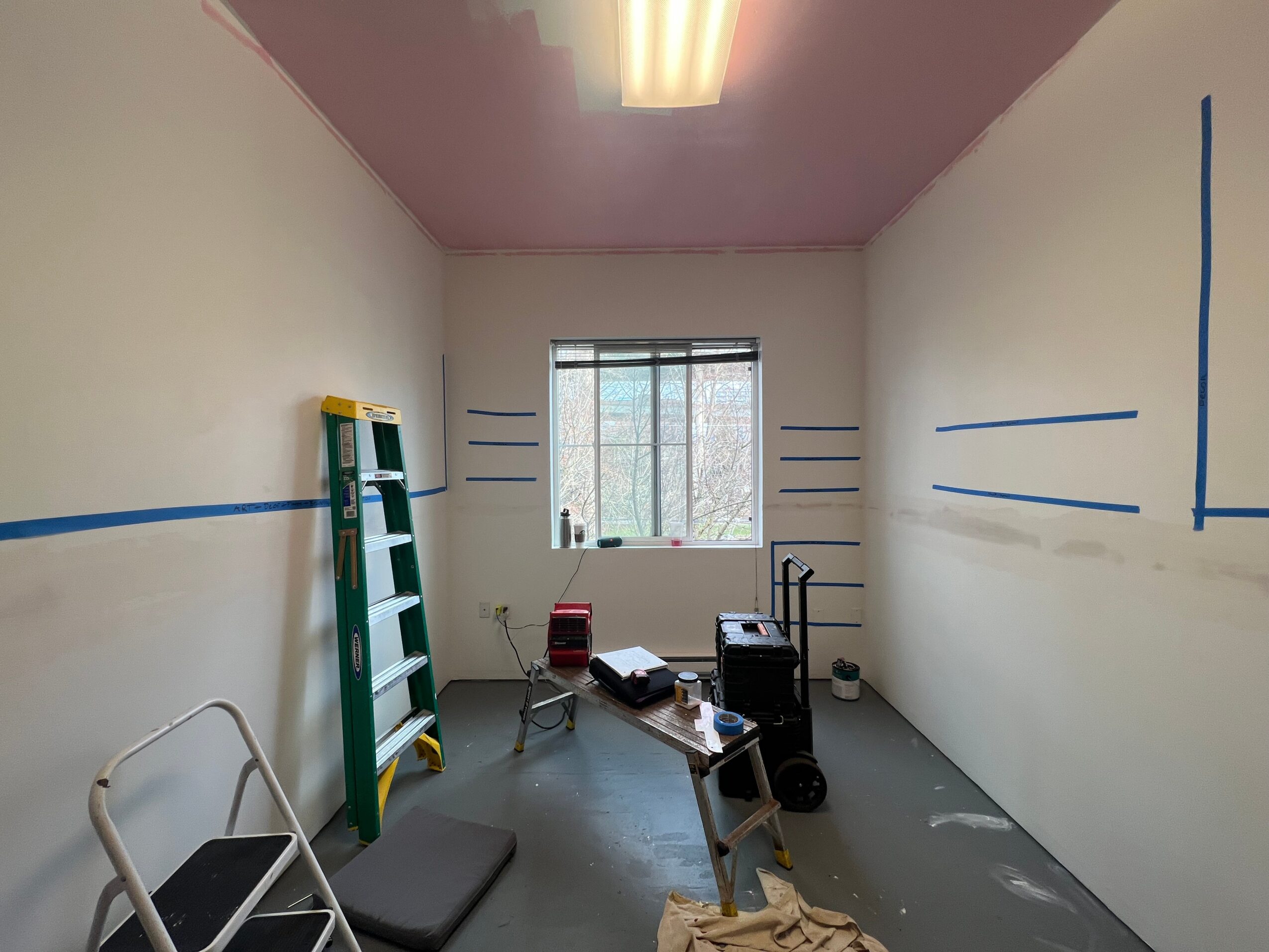 Empty room with white walls, a window, a ladder, and some equipment, painted with blue lines, ceiling light, and gray floor.