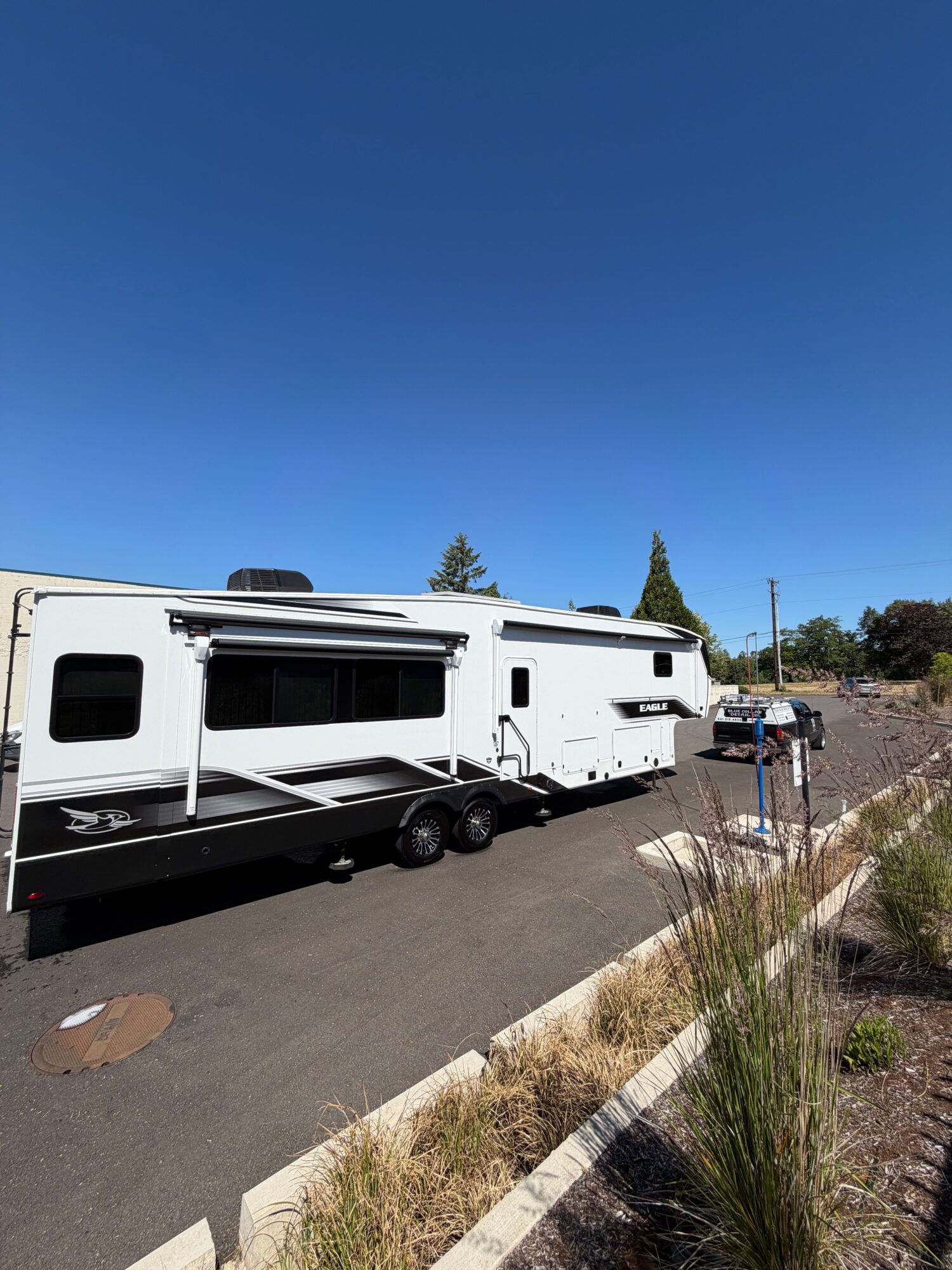 A white RV parked on a paved road with a clear blue sky and trees in the background.