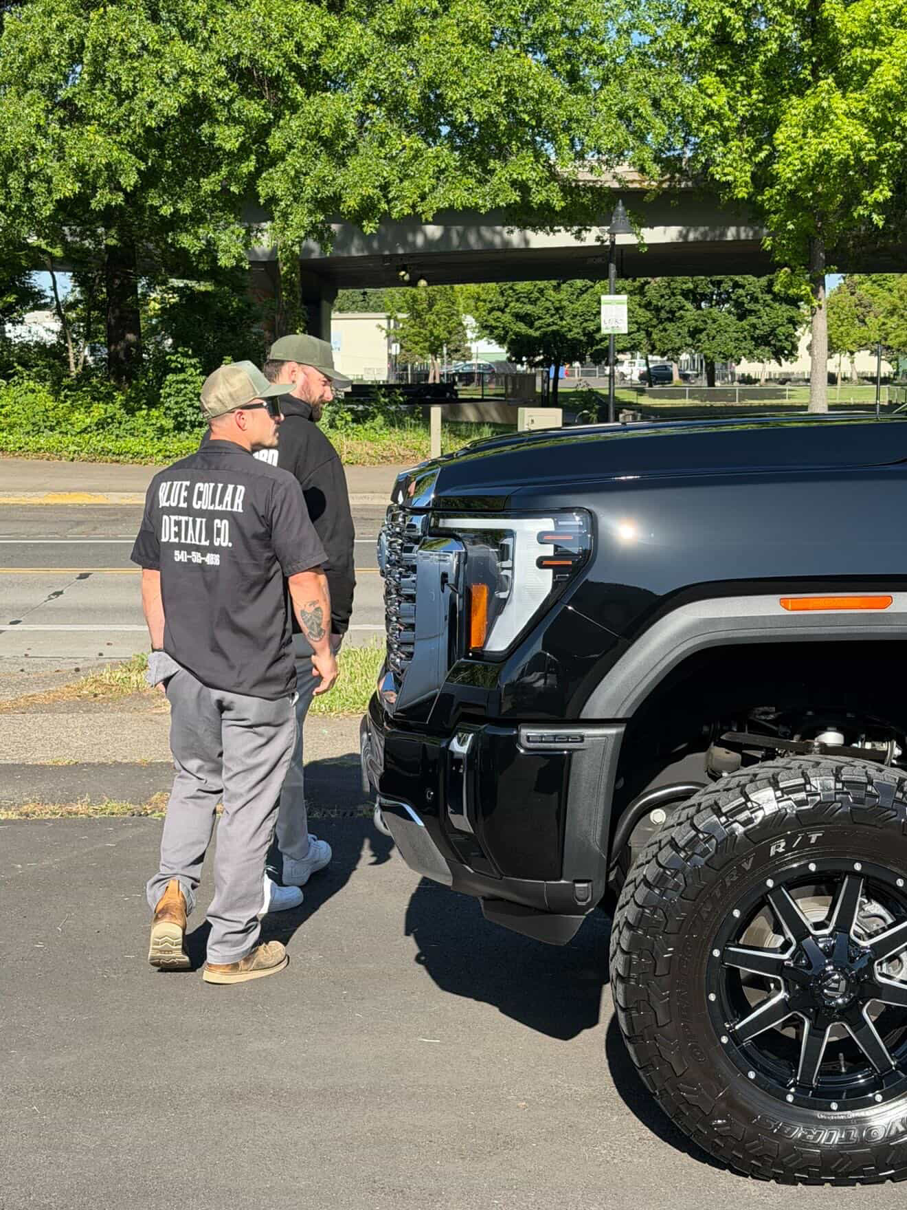 Two men stand near a black pickup truck on a street with trees and a bridge in the background.