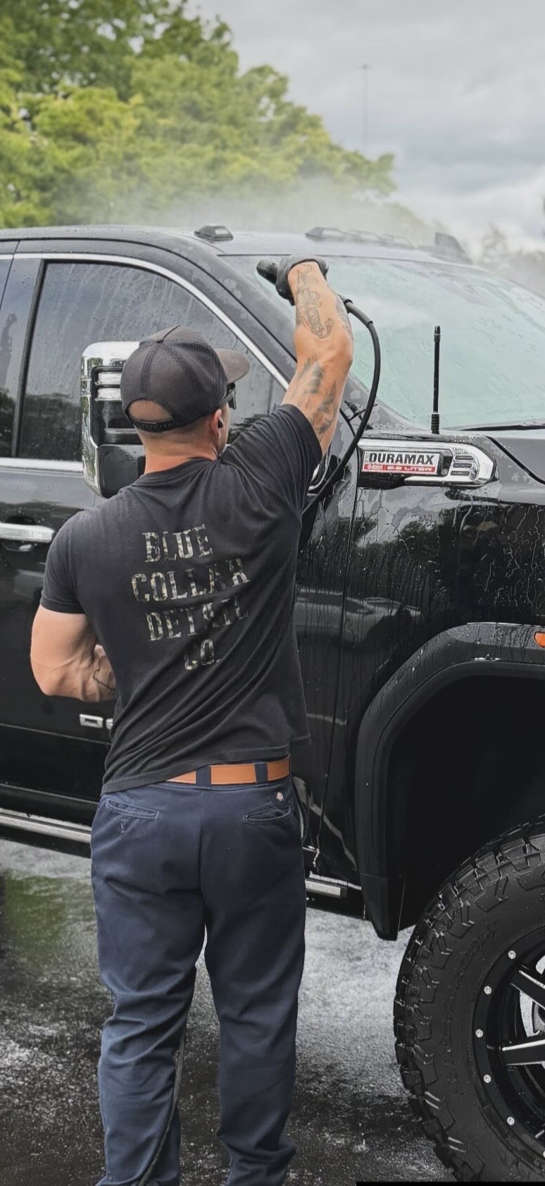 Person cleaning a black pickup truck outdoors, with trees and cloudy sky in background.