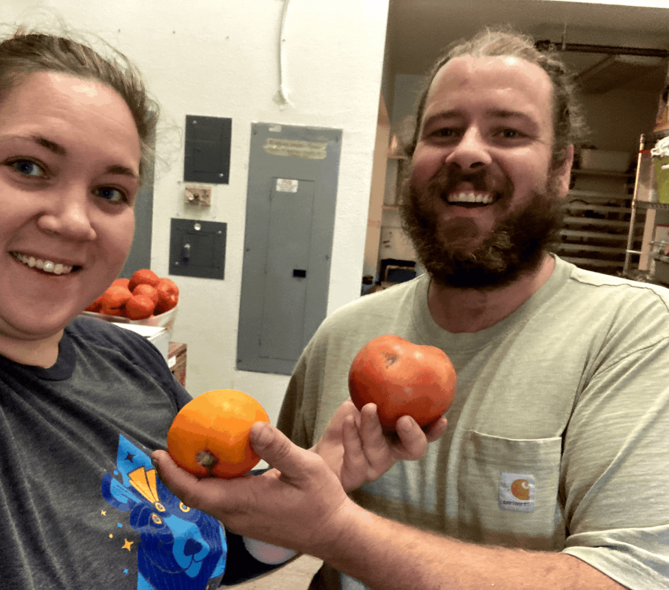 Two people smiling indoors, each holding a fruit, with electrical panels in the background.
