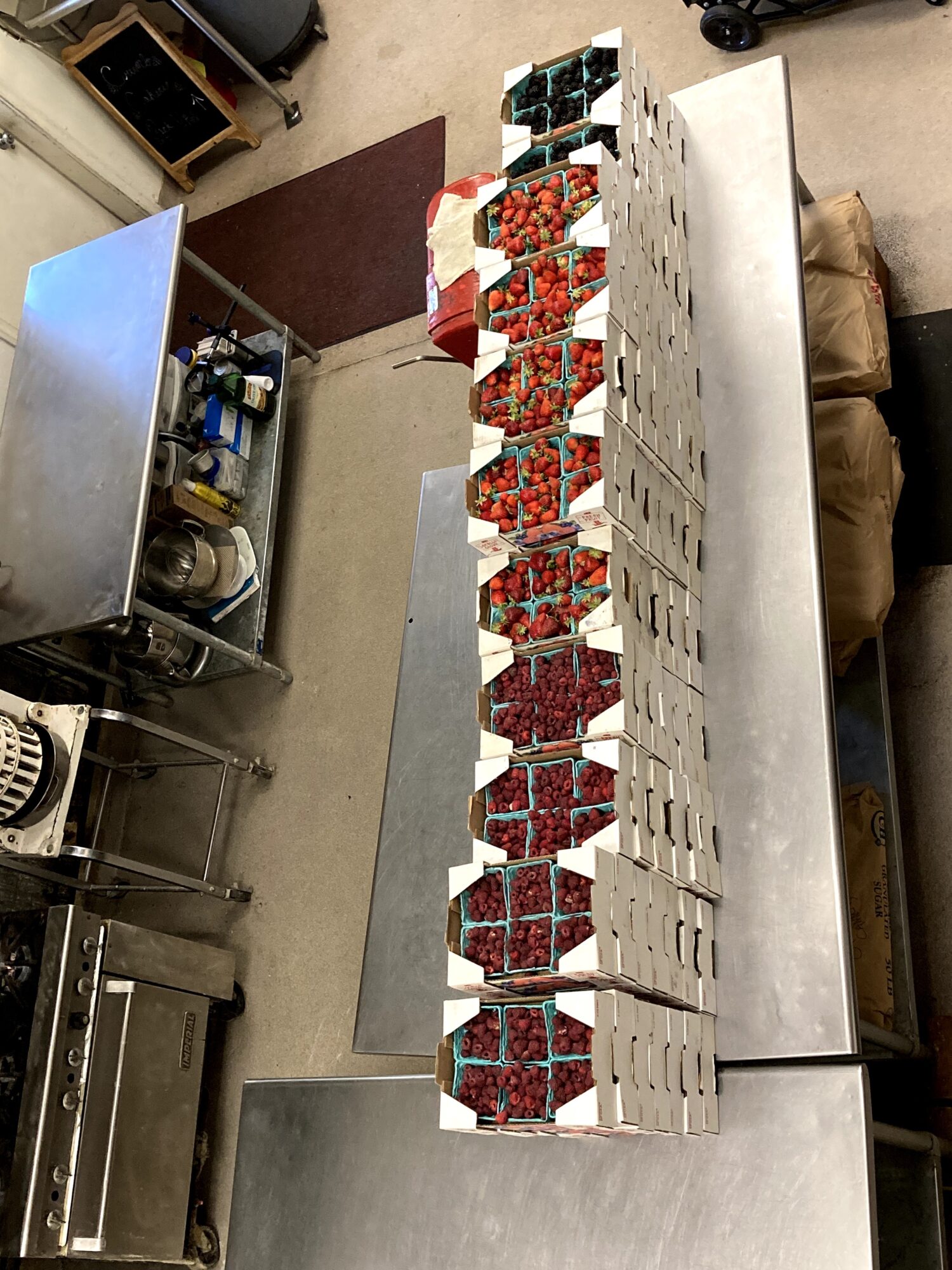 View of a long table with stacked boxes containing red and blue items, in a room with kitchen equipment and furniture.