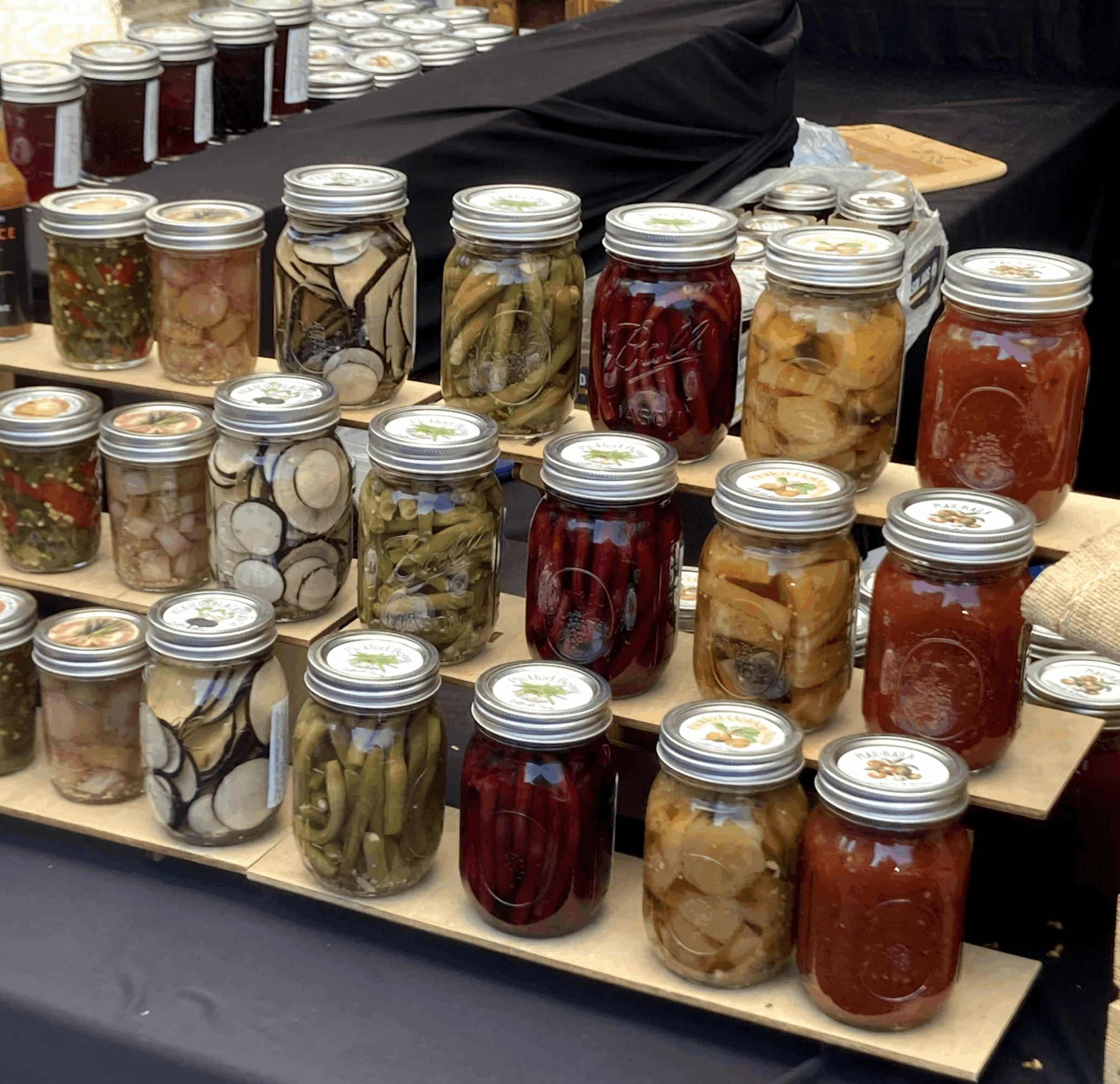 Display of various jars filled with preserved vegetables and fruits on a table at a market or store.