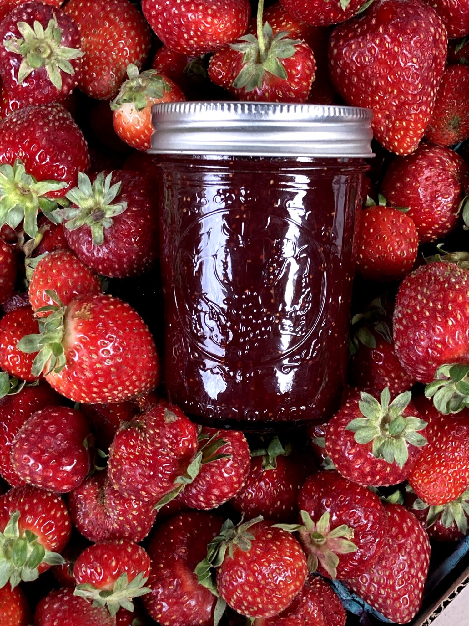 Jar of strawberry jam surrounded by fresh strawberries, some with green leaves, on a surface.